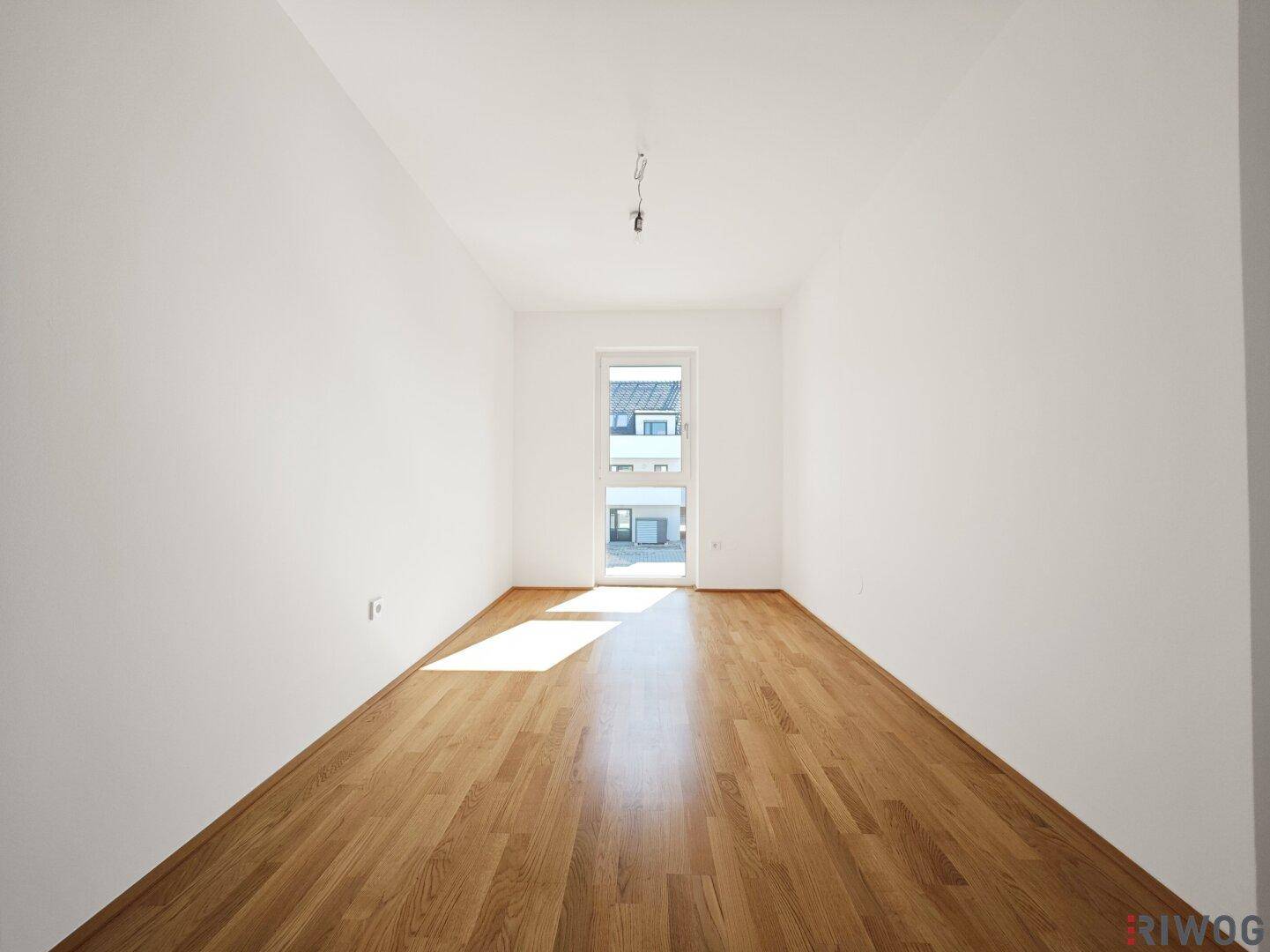 Leeres, sonniges Zimmer mit Parkettboden und Zugang zu einem französischen Balkon.