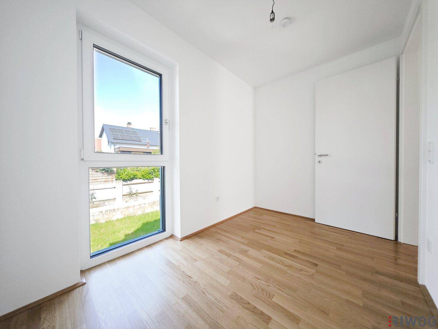 Helles Zimmer mit Parkettboden und großem Fenster mit Blick auf den grünen Garten.