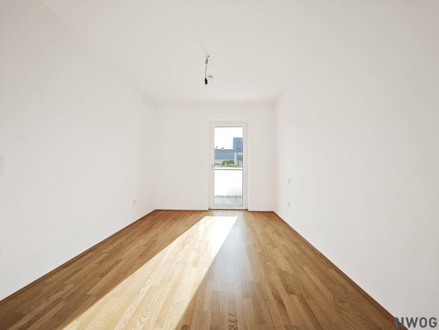 Lichtdurchflutetes Zimmer mit Holzboden und Tür zu einem kleinen Balkon oder Austritt.