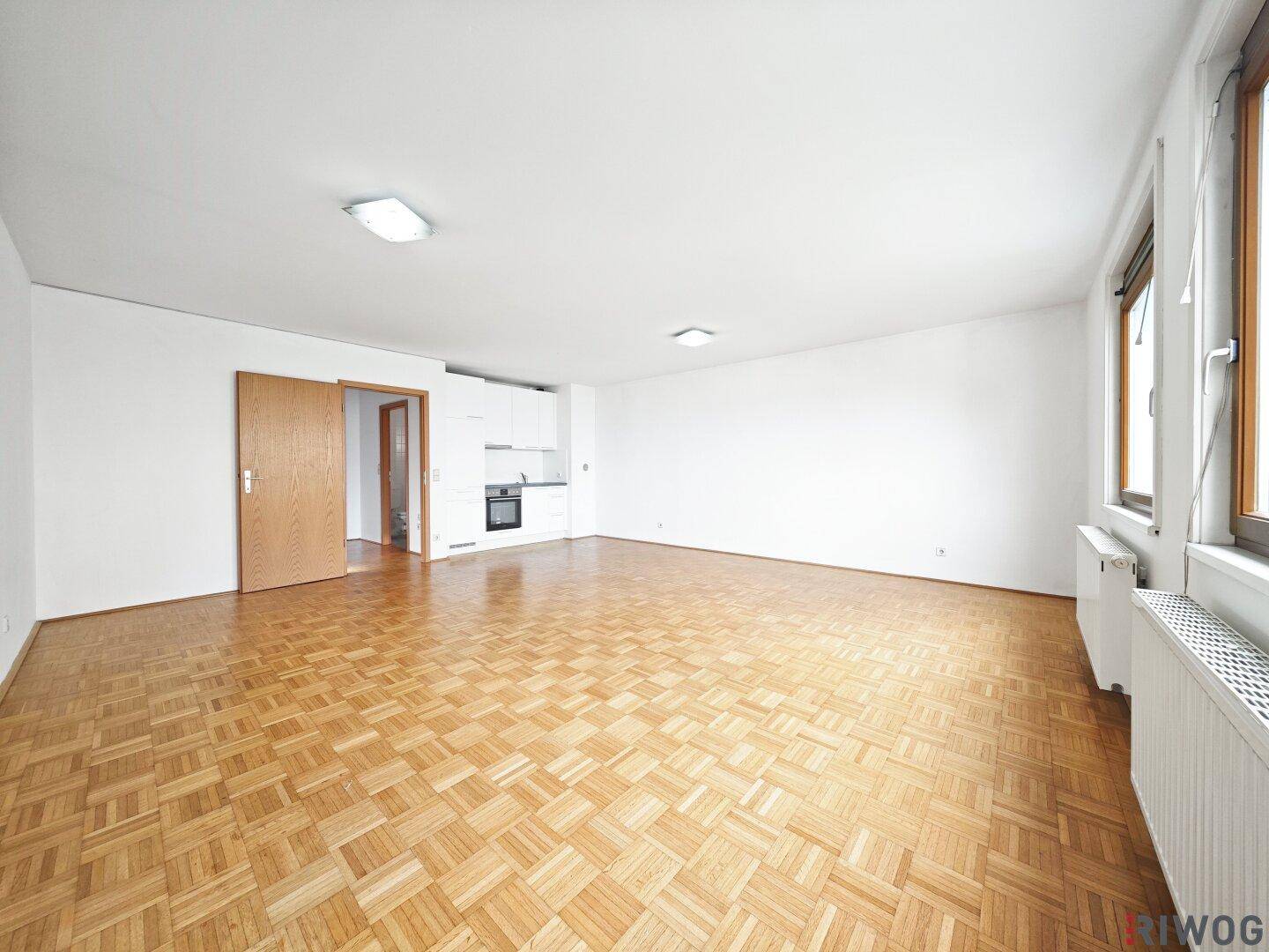 Geräumiger Wohnbereich mit Parkettboden und integrierter Küchenzeile, lichtdurchflutet durch Fenster.