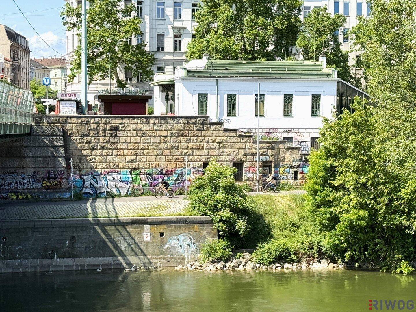 Gebäude am Flussufer mit historischer Steinmauer und viel Grün in der Umgebung.