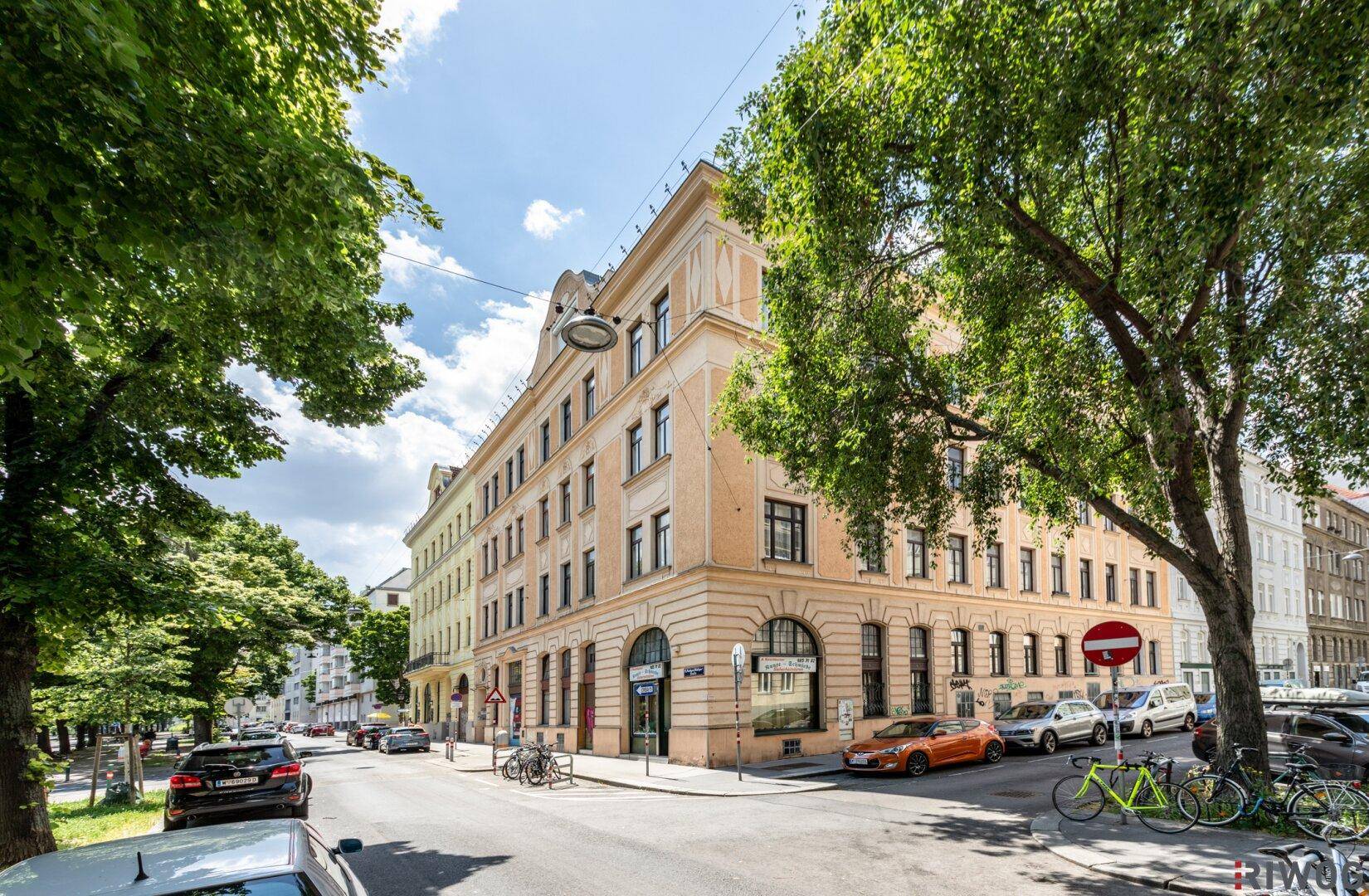 Eckgebäude in einer städtischen Umgebung mit Bäumen und geparkten Fahrzeugen an einem sonnigen Tag.