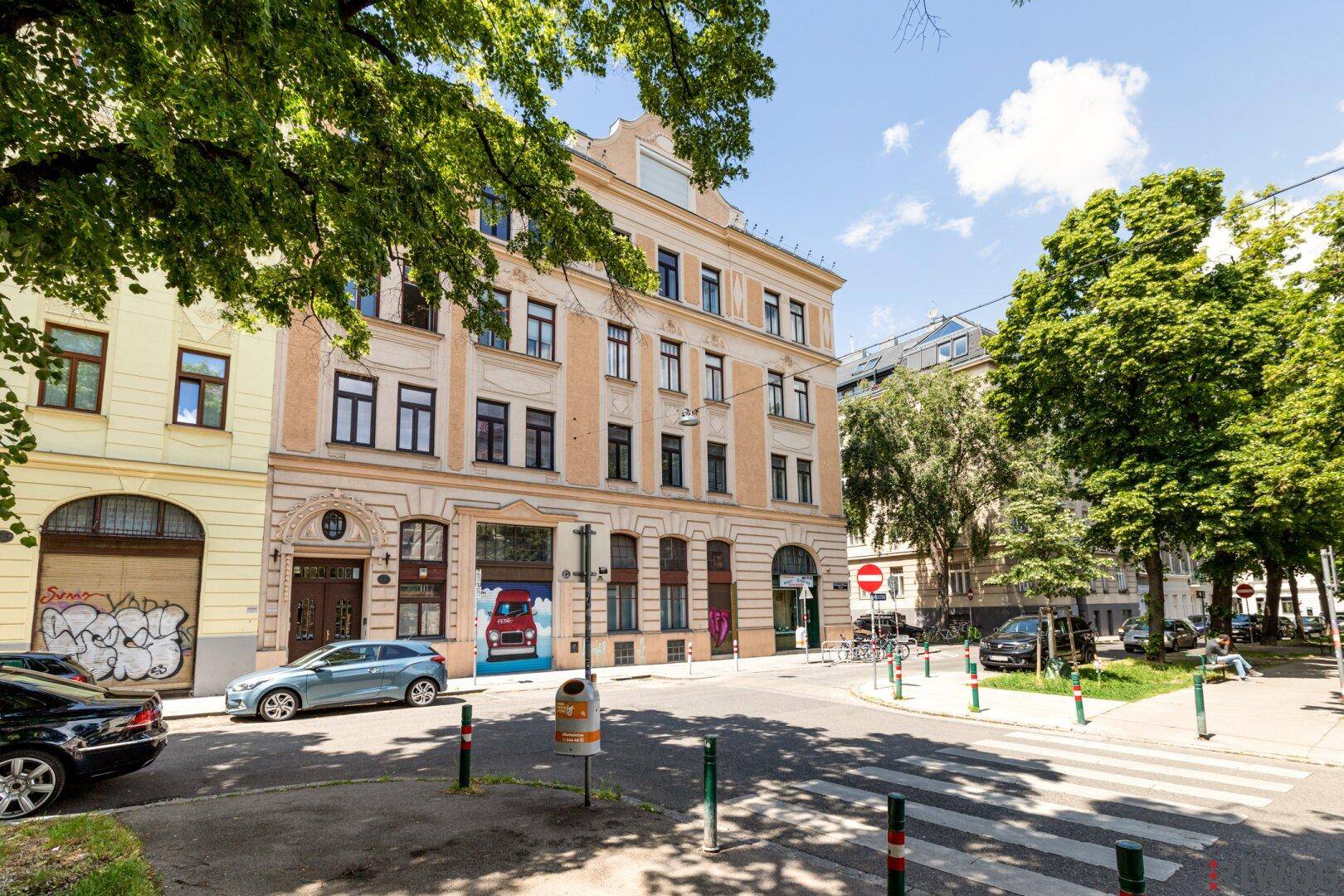 Historisches Wohngebäude an einer belebten Straße mit Bäumen und parkenden Autos.