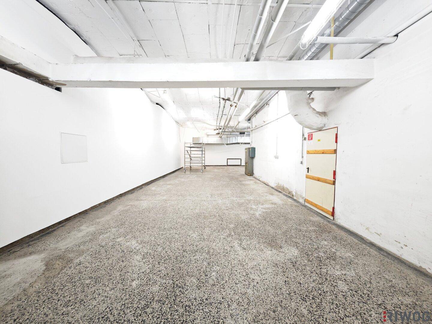 Geräumiger Kellerraum mit Betonboden, freiliegenden Deckenstrukturen und einer Brandschutztür.