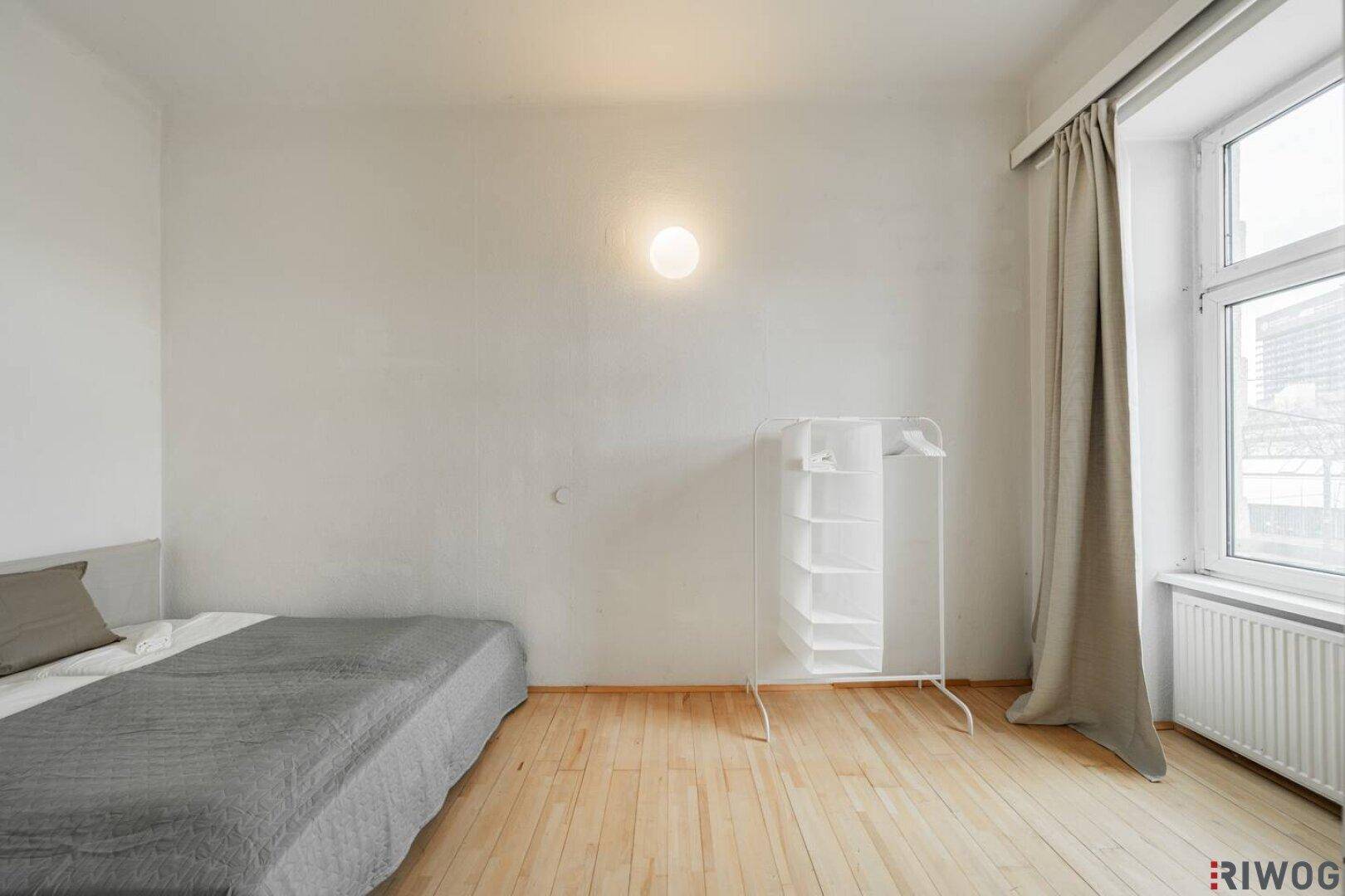 Schlichtes Schlafzimmer mit Holzboden, großem Fenster und praktischem Kleiderständer.