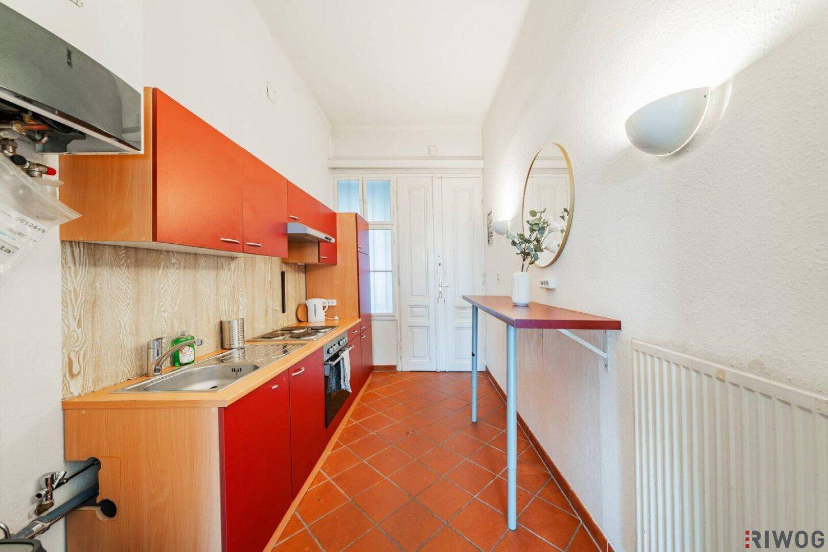 Funktionale Küche mit roten Schränken, Terrakotta-Fliesen und einem Heizkörper unter dem Fenster.