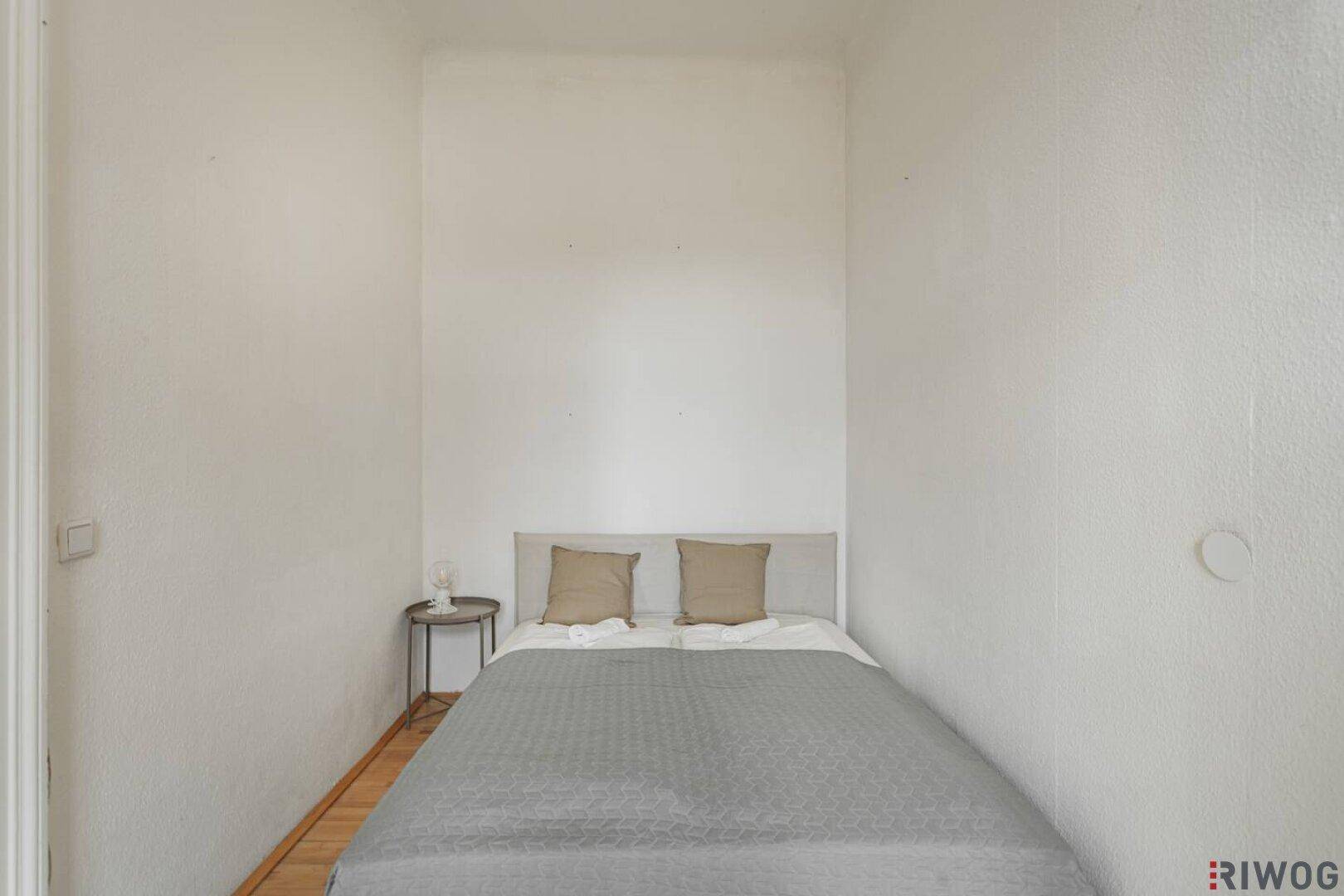 Kleines, helles Schlafzimmer mit Einzelbett, Nachttisch und schlichtem Holzboden.