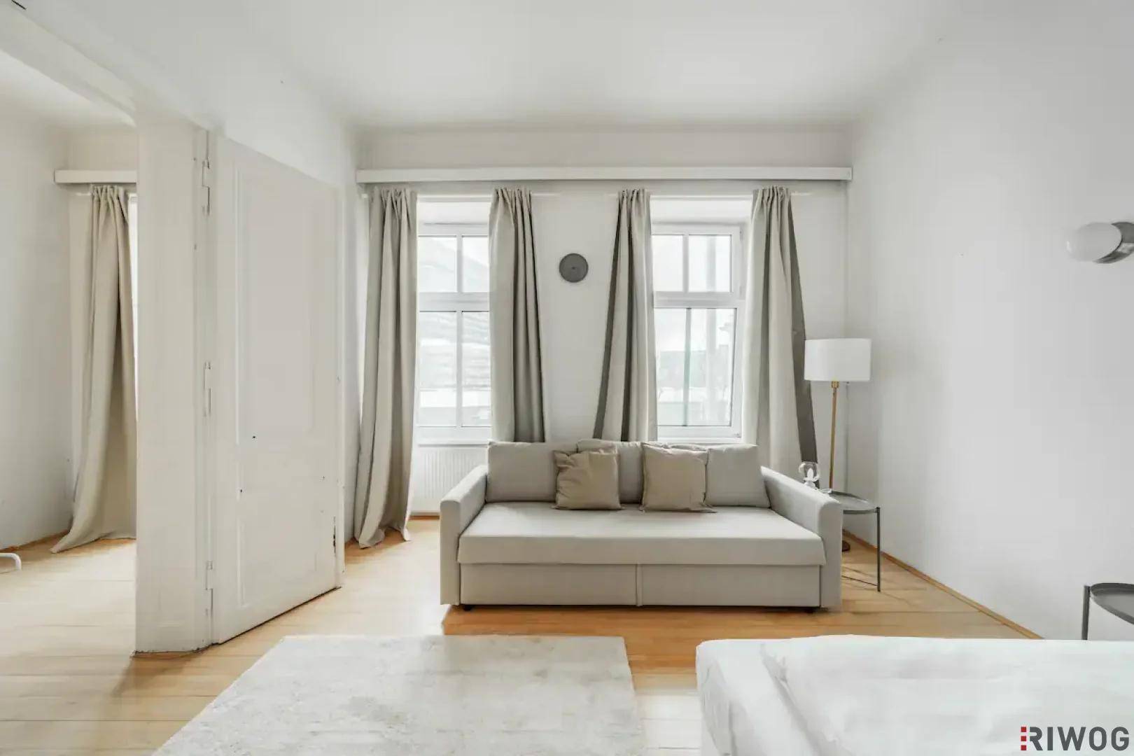 Helles Schlafzimmer mit Holzboden, einem Schlafsofa und großen Fenstern mit hellen Vorhängen.
