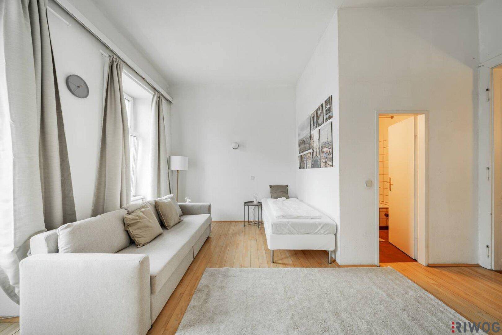 Heller Wohnbereich mit Sofa, Einzelbett und Holzboden, der zu einem Badezimmer führt.