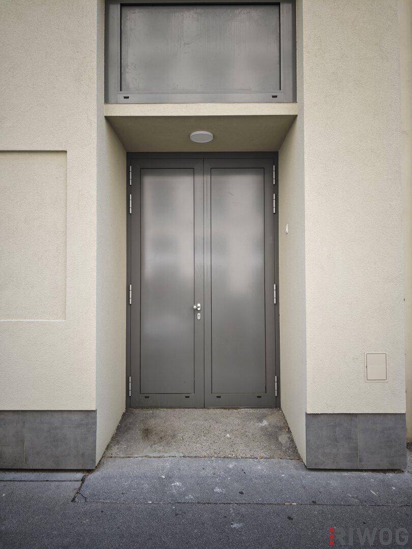 Graue Doppeltür als Eingang zu einem Gebäude mit kleiner Rampe und Überdachung.