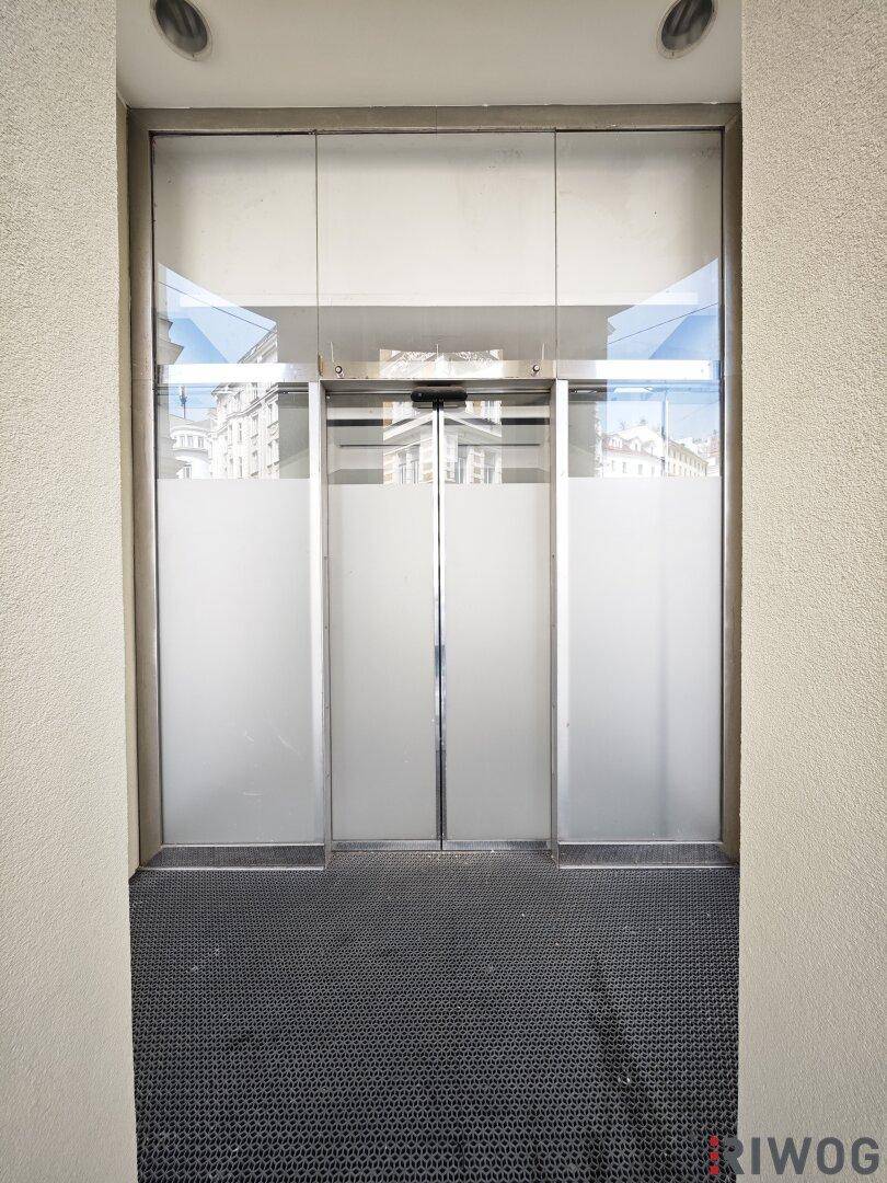 Eingangsbereich mit automatischen Glastüren und dunklem Teppichboden, der nach außen führt.