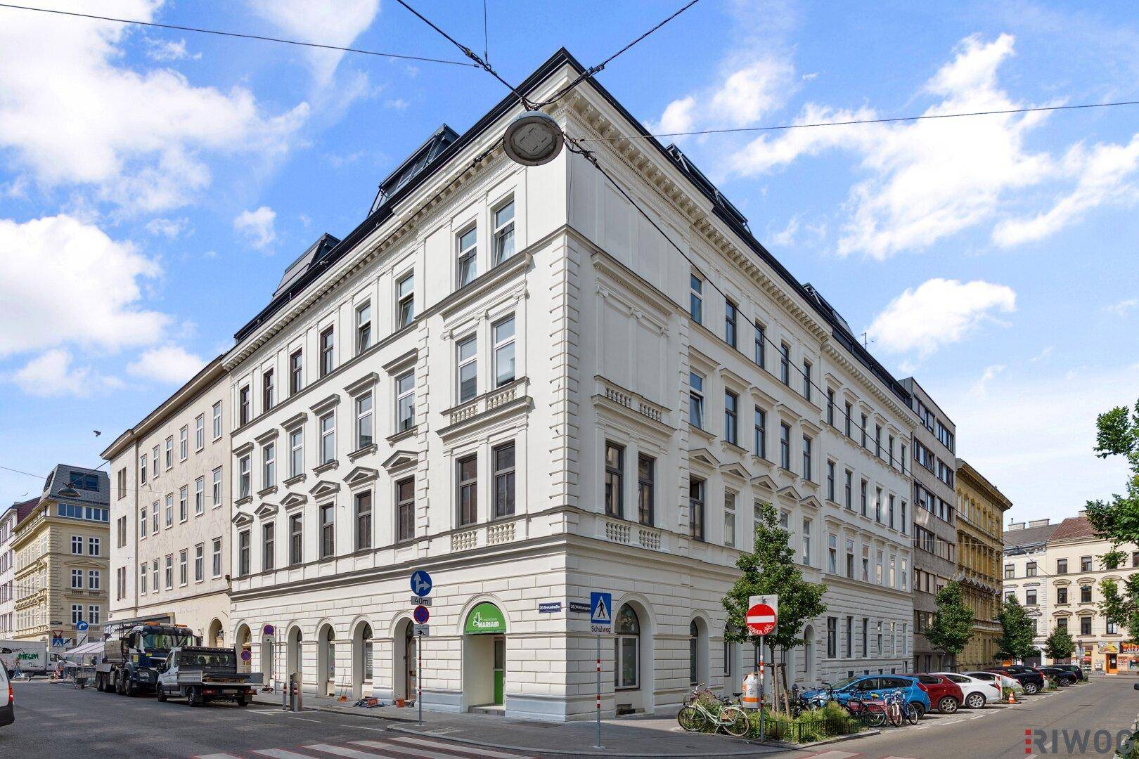 Weißes Eckgebäude mit klassischer Architektur, vielen Fenstern und blauem Himmel.
