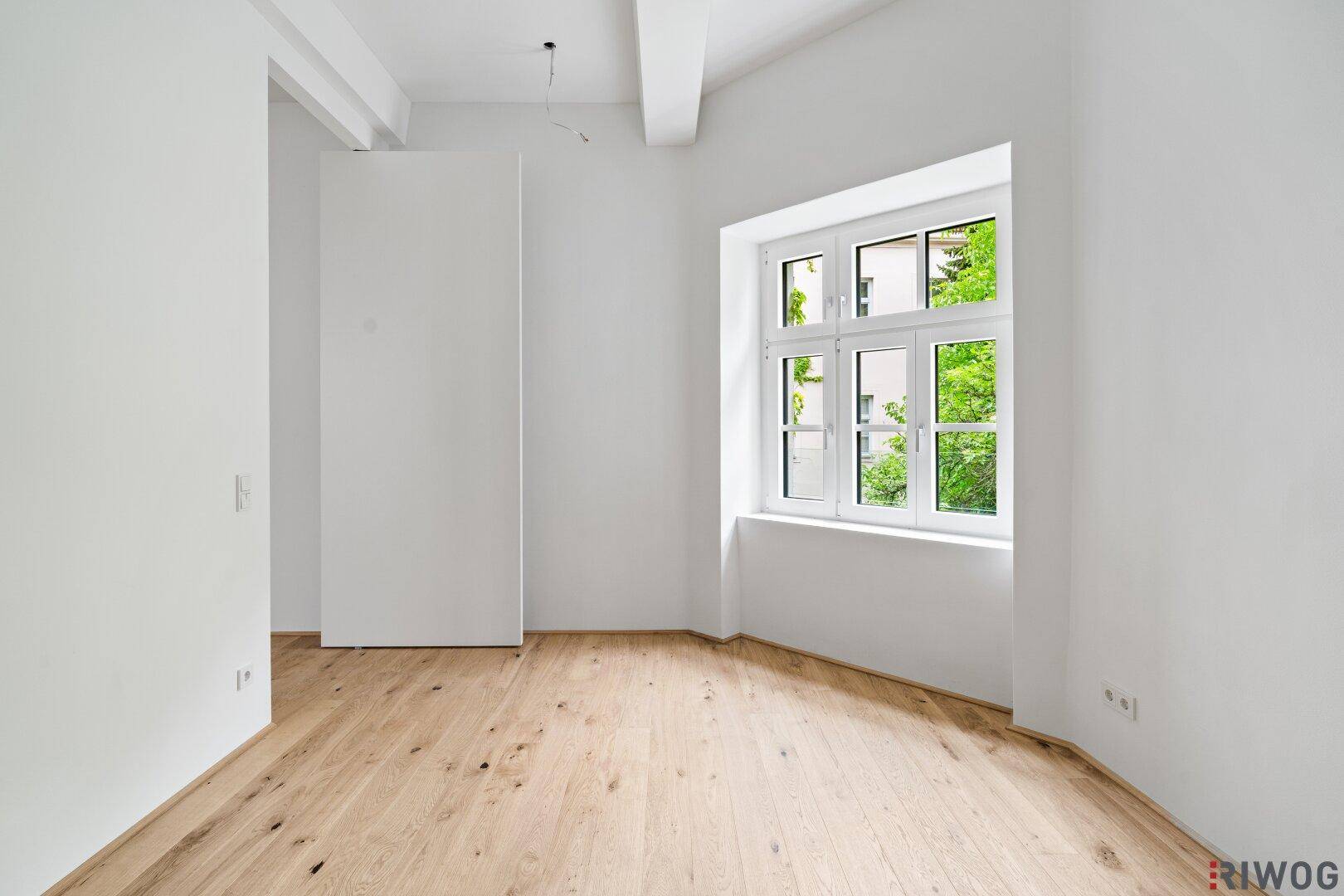 Helles Zimmer mit Holzboden und einem großen Fenster mit Blick ins Grüne.