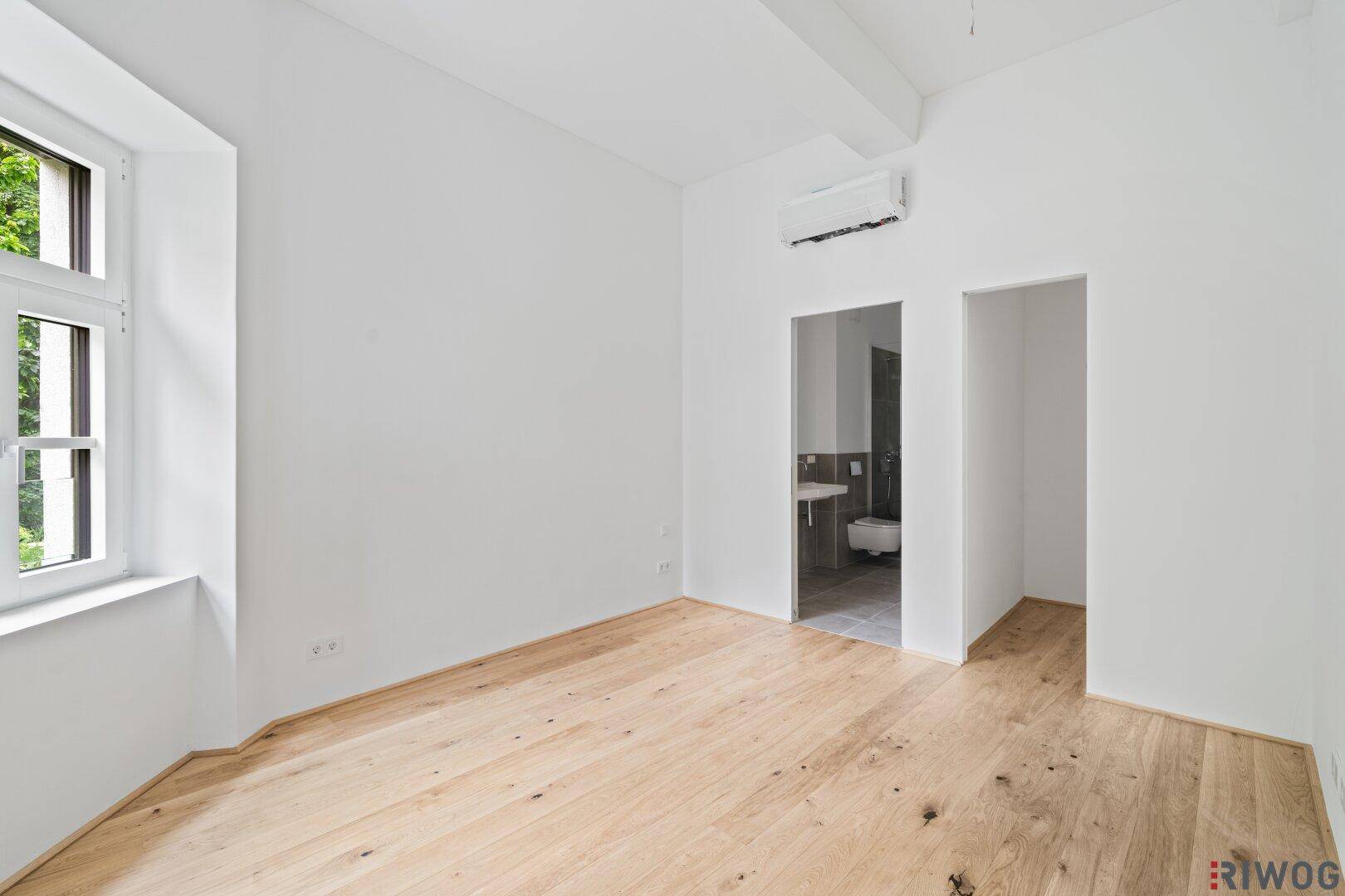 Helles Schlafzimmer mit Holzboden, Fensterblick ins Grüne und Zugang zu einem Badezimmer.