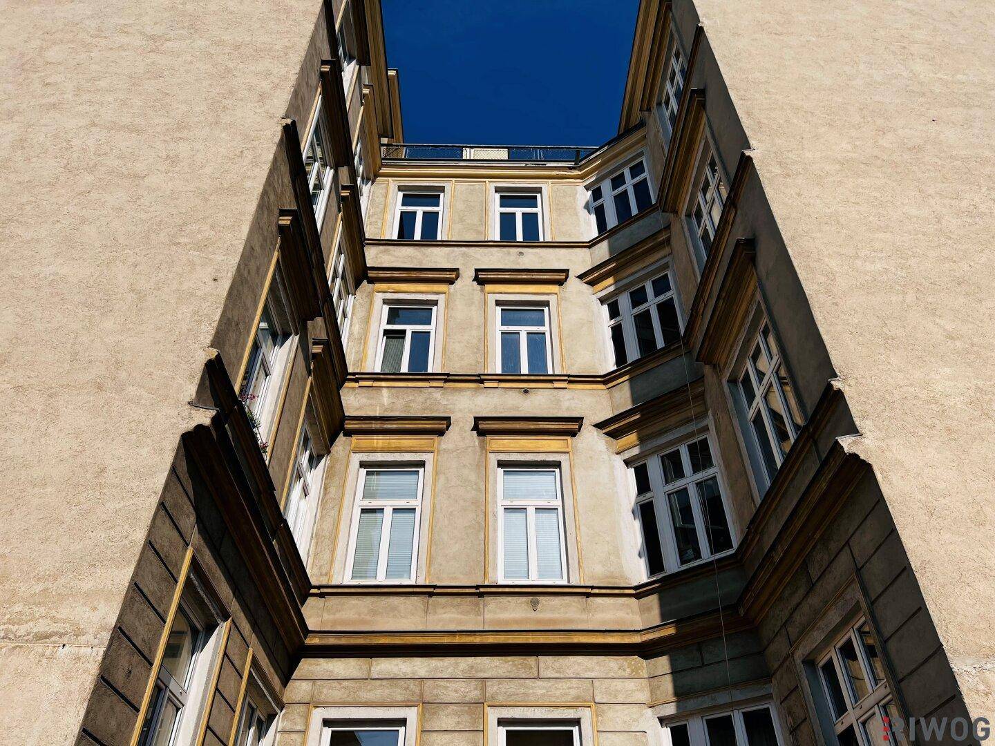 Blick in den Innenhof eines Altbaus mit mehreren Etagen und vielen Fenstern unter blauem Himmel.