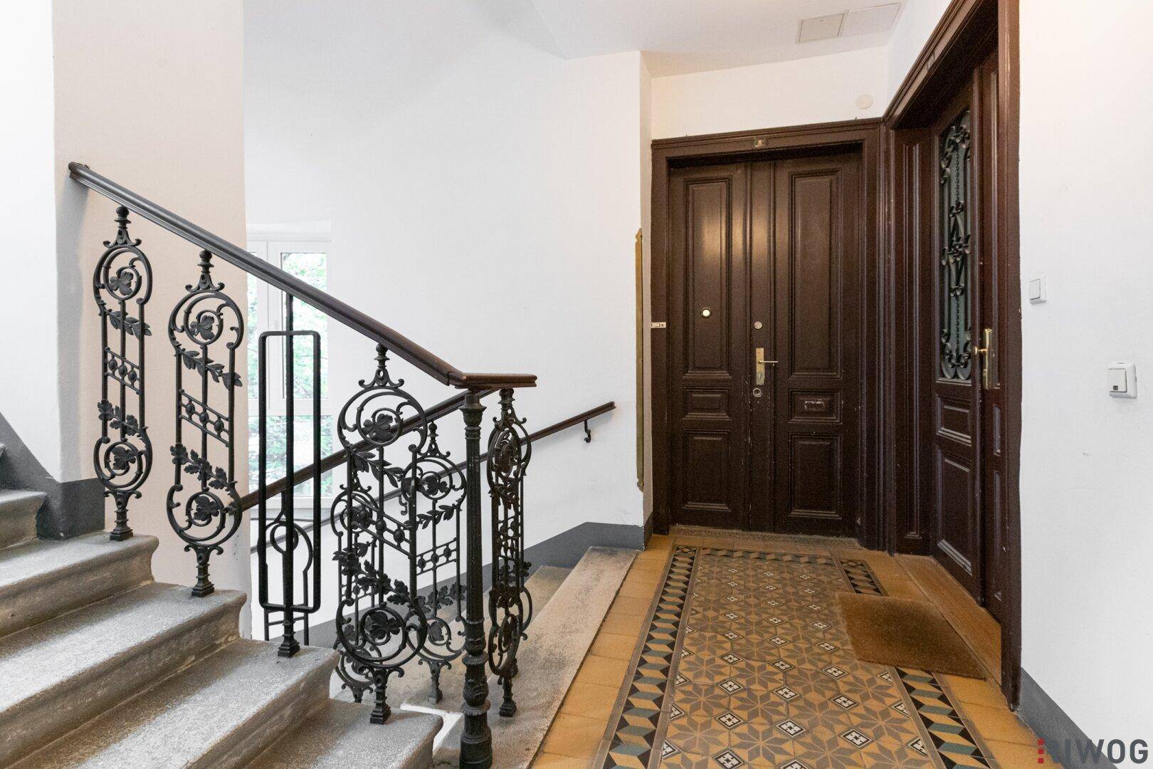 Historisches Treppenhaus mit schmiedeeisernem Geländer und gemustertem Fliesenboden im Eingangsbereich.