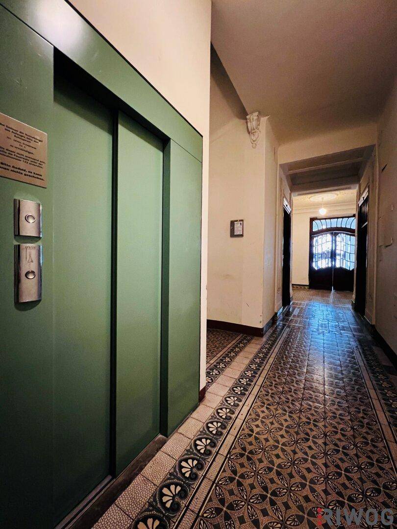 Langer Flur in einem Altbau mit gemusterten Fliesen und einem grünen Aufzug.