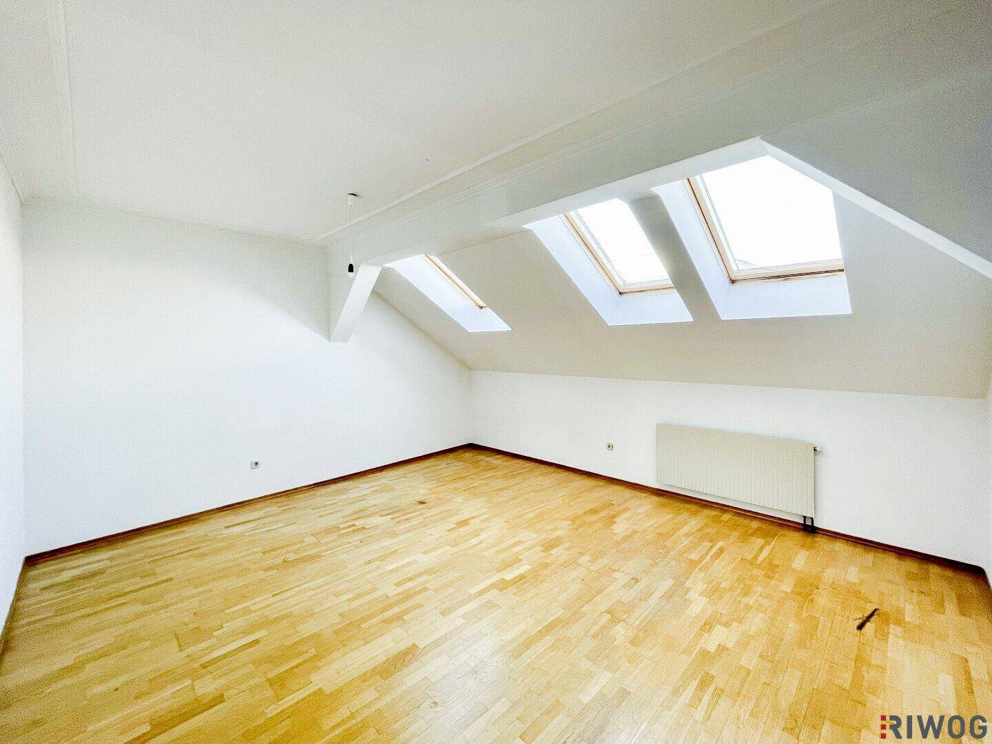 Geräumiges Dachzimmer mit schrägen Wänden, Holzboden und mehreren Dachfenstern für viel natürliches Licht.