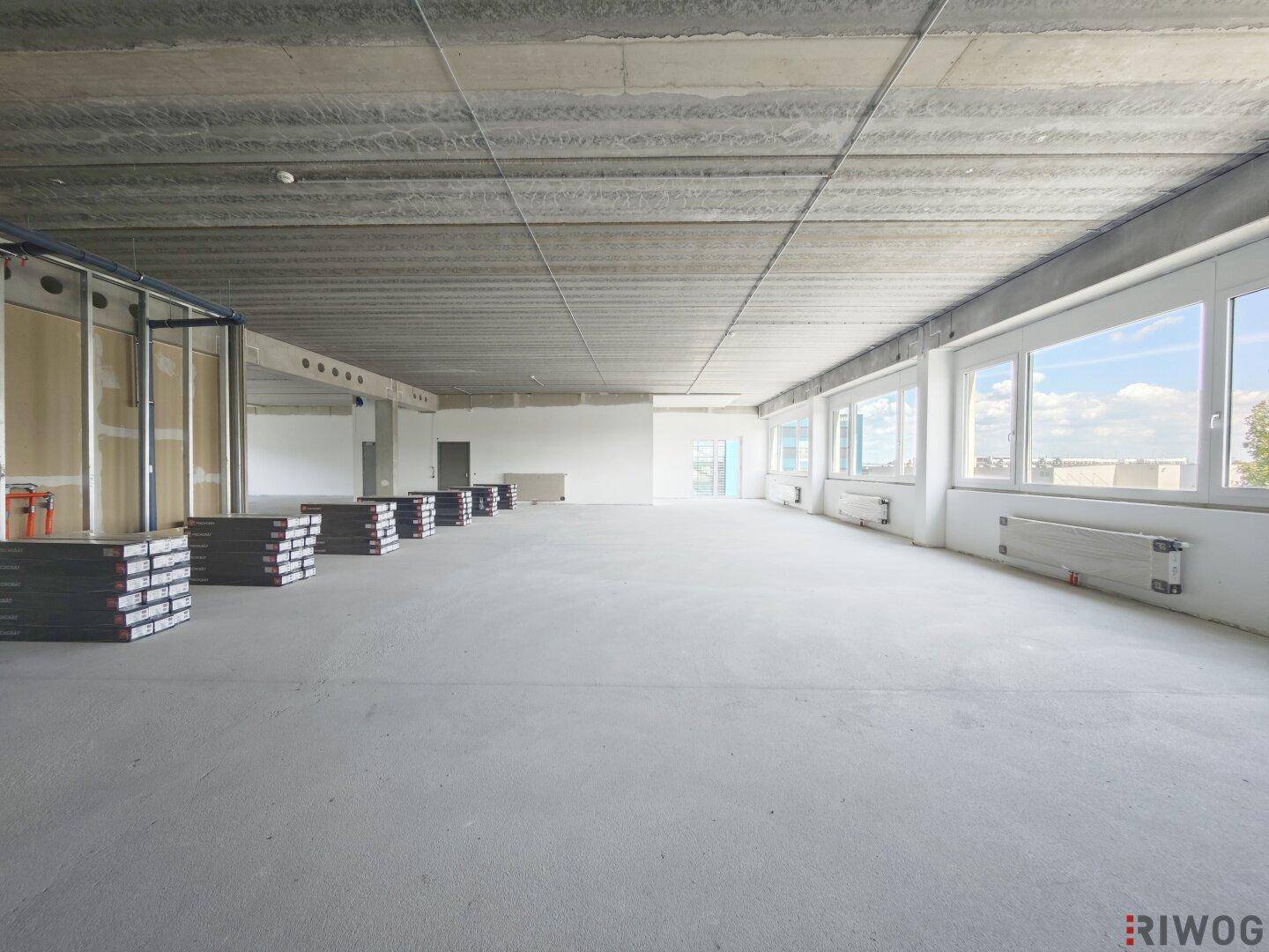 Großer, heller Büroraum im Rohbau mit vielen Fenstern und einer markanten Betondecke, bereit zur Fertigstellung.