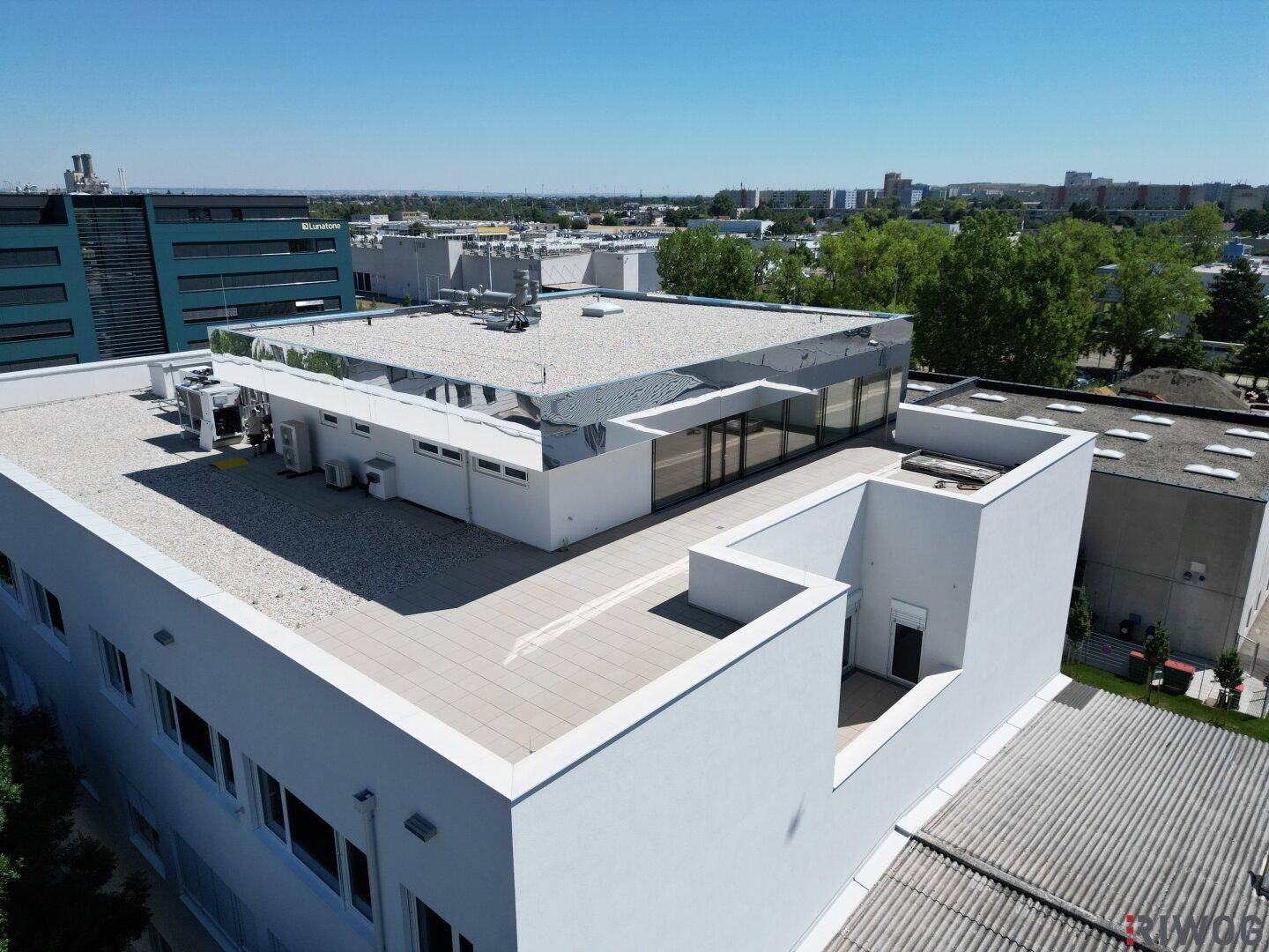 Dachterrasse mit Kiesbelag und Klimaanlagen, umgeben von weiteren Gebäuden und Grünflächen.