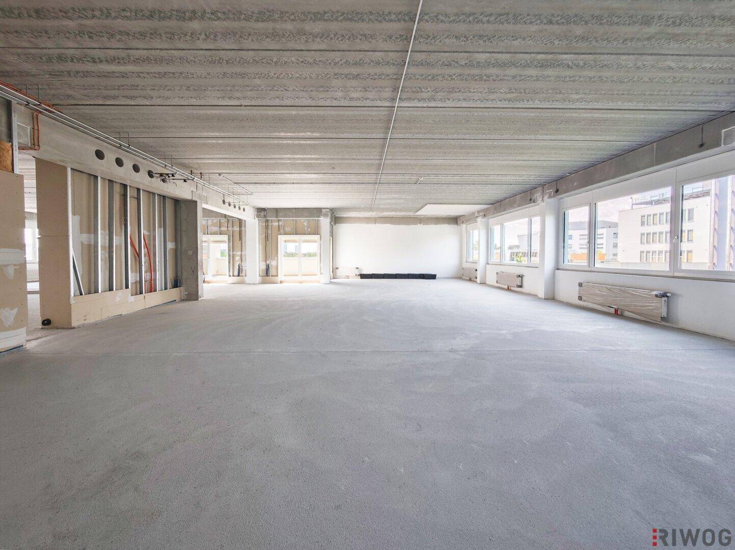 Geräumiger Büroraum im Rohbau mit vielen Fenstern und Blick auf die Umgebung.