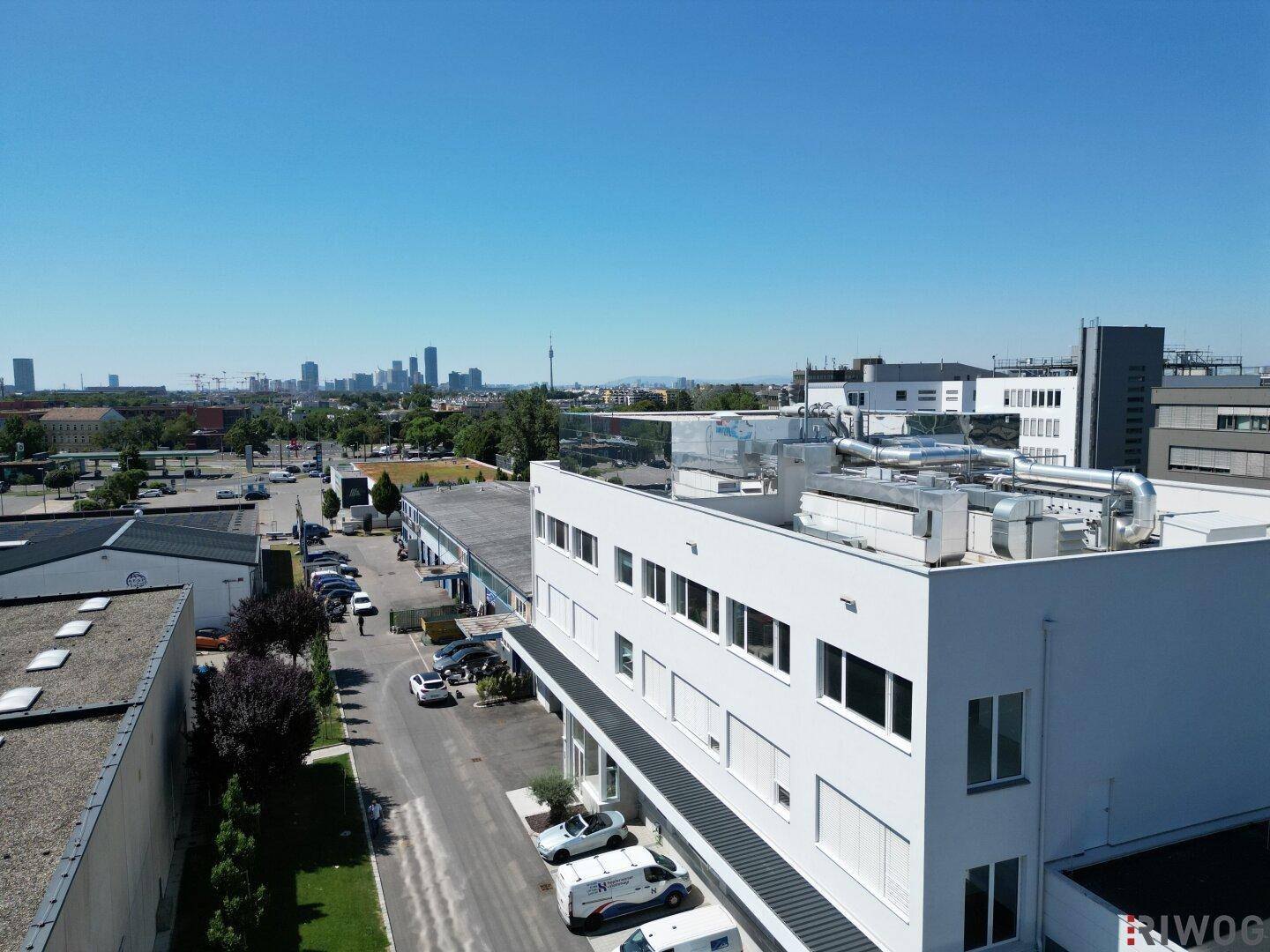 Luftaufnahme eines modernen Bürogebäudes mit Parkplätzen und Stadtpanorama im Hintergrund.