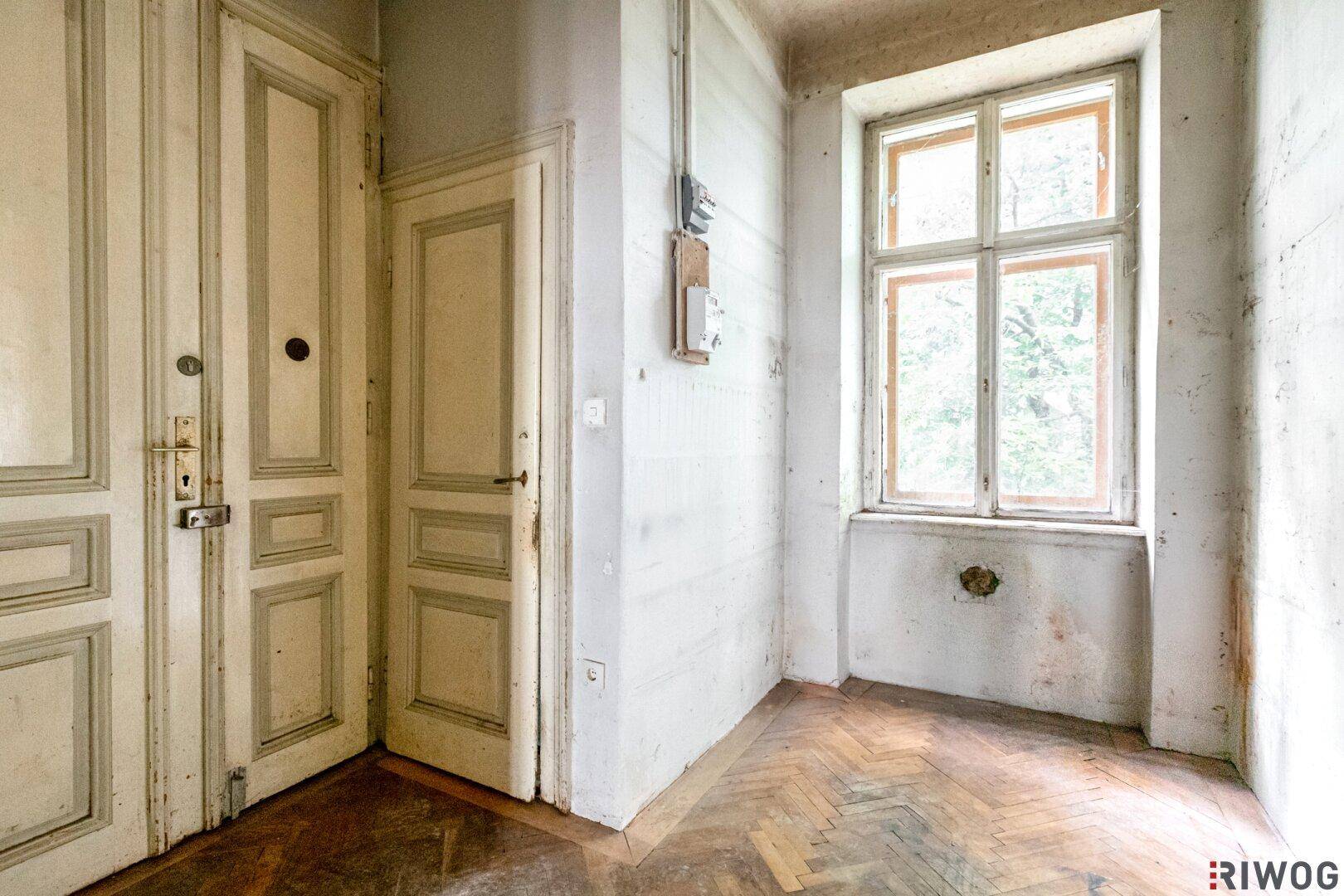 Leerer Raum mit abgenutztem Fischgrätparkett und alten Holztüren, bereit zur Sanierung.