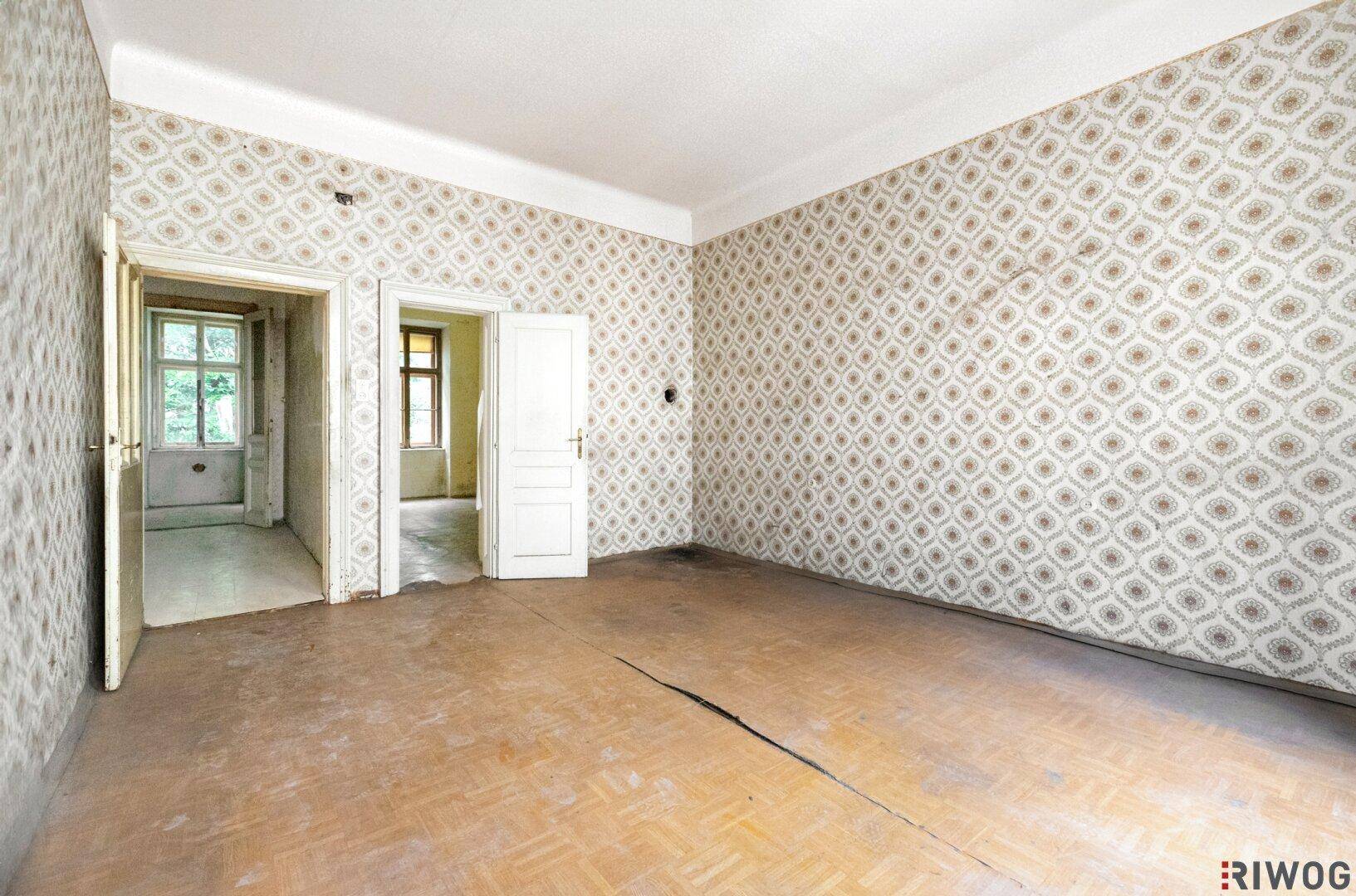 Leerer Raum mit alter Tapete, Holzboden und zwei offenen Türen zu weiteren Zimmern.