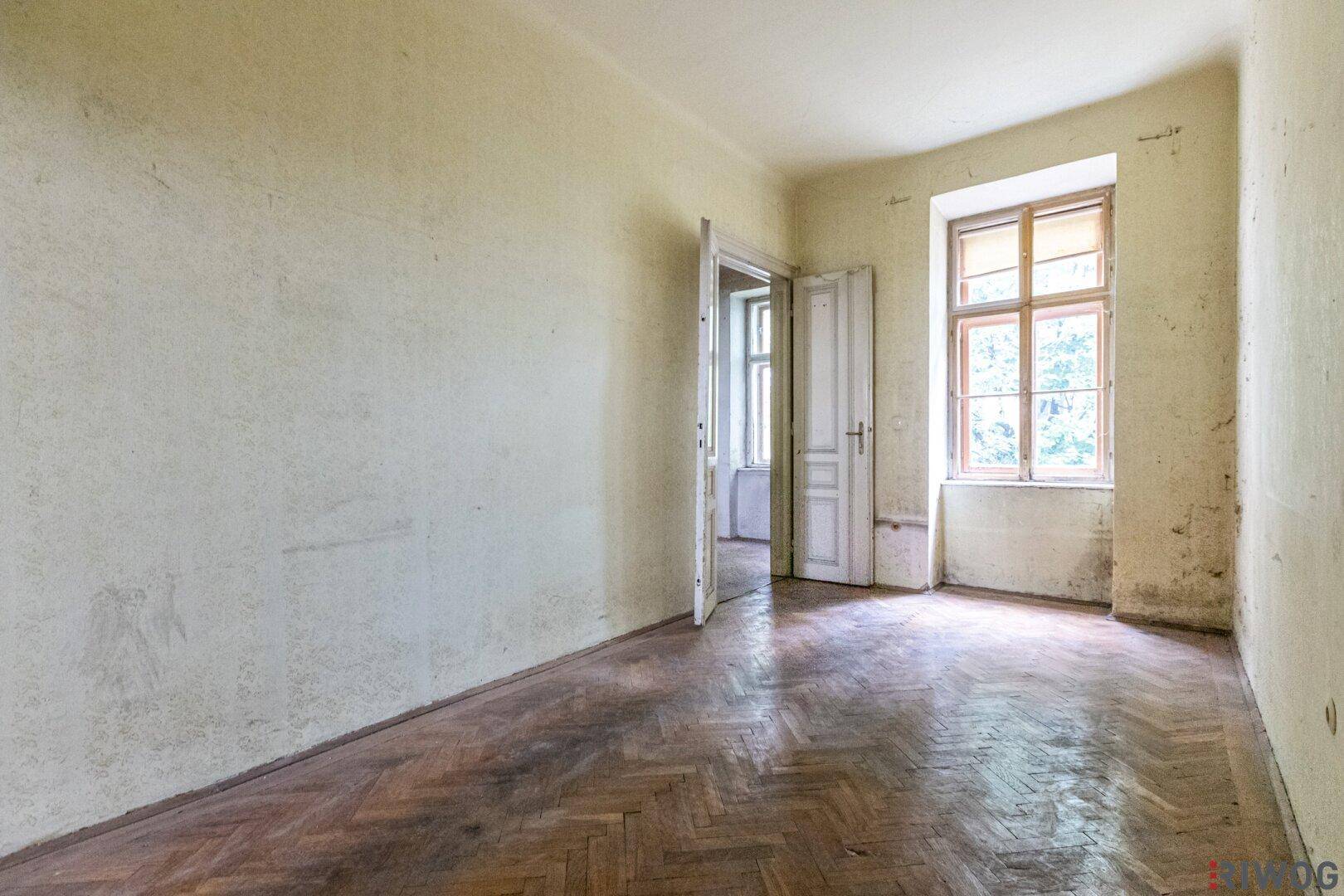 Leerer Raum mit abgenutztem Fischgrätparkett und renovierungsbedürftigen Wänden, bereit zur Sanierung.