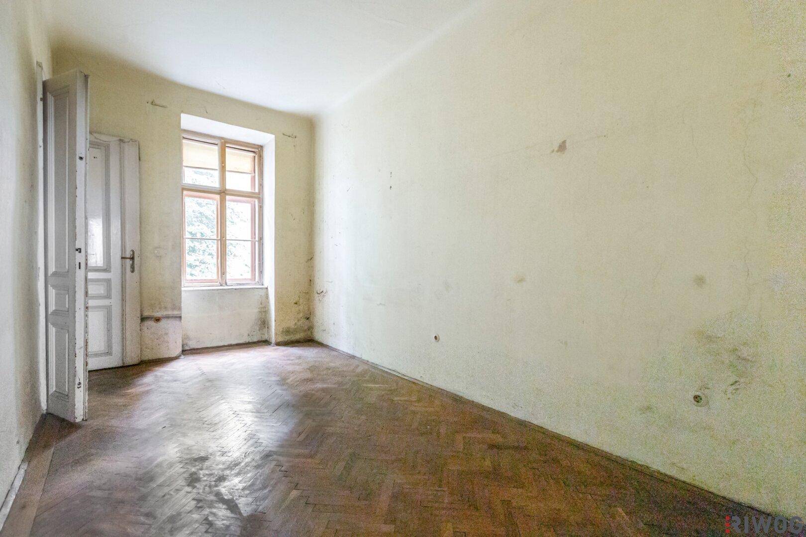 Leerer Raum mit abgenutztem Fischgrätparkett und renovierungsbedürftigen Wänden, bereit zur Sanierung.