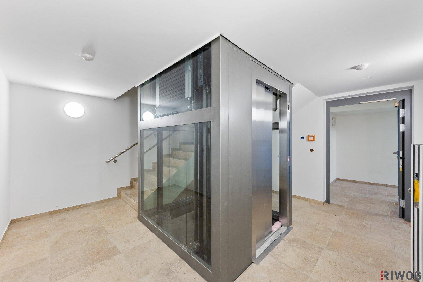 Moderner Aufzug in einem hellen Treppenhaus mit Fliesenboden.