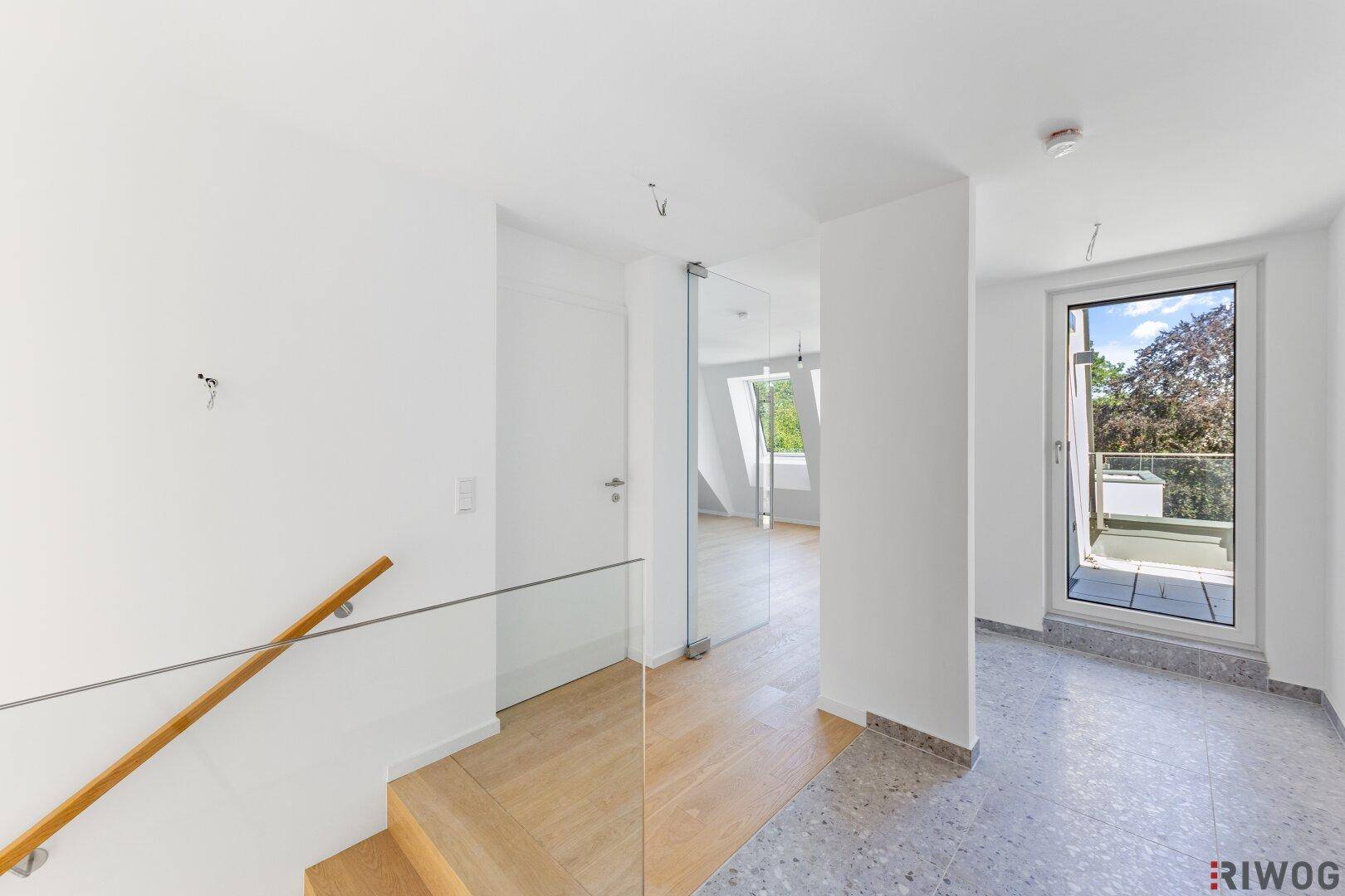 Heller Flur mit Holztreppe, Glasgeländer und Zugang zu weiteren Räumen und Balkon.