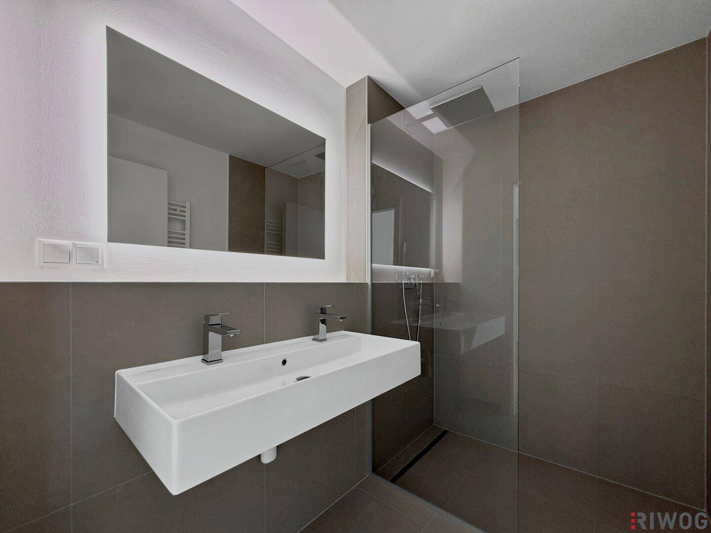 Elegantes Badezimmer mit Doppelwaschbecken, beleuchtetem Spiegel und geräumiger ebenerdiger Dusche.