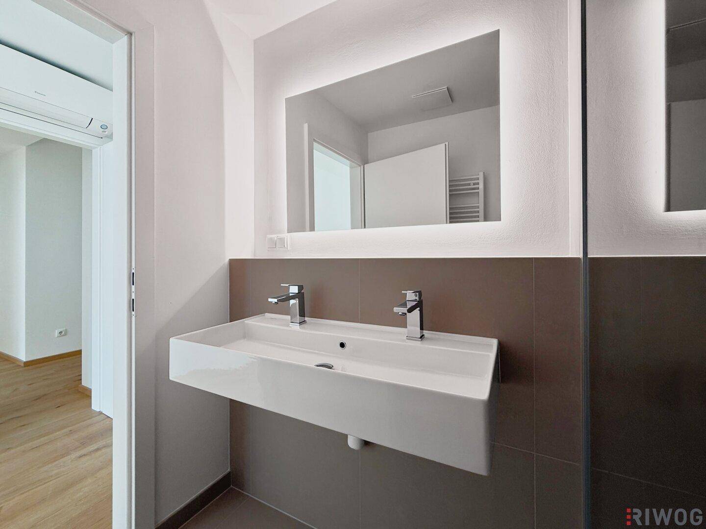 Modernes Badezimmer mit Doppelwaschbecken, beleuchtetem Spiegel und Zugang zu einem weiteren Raum.
