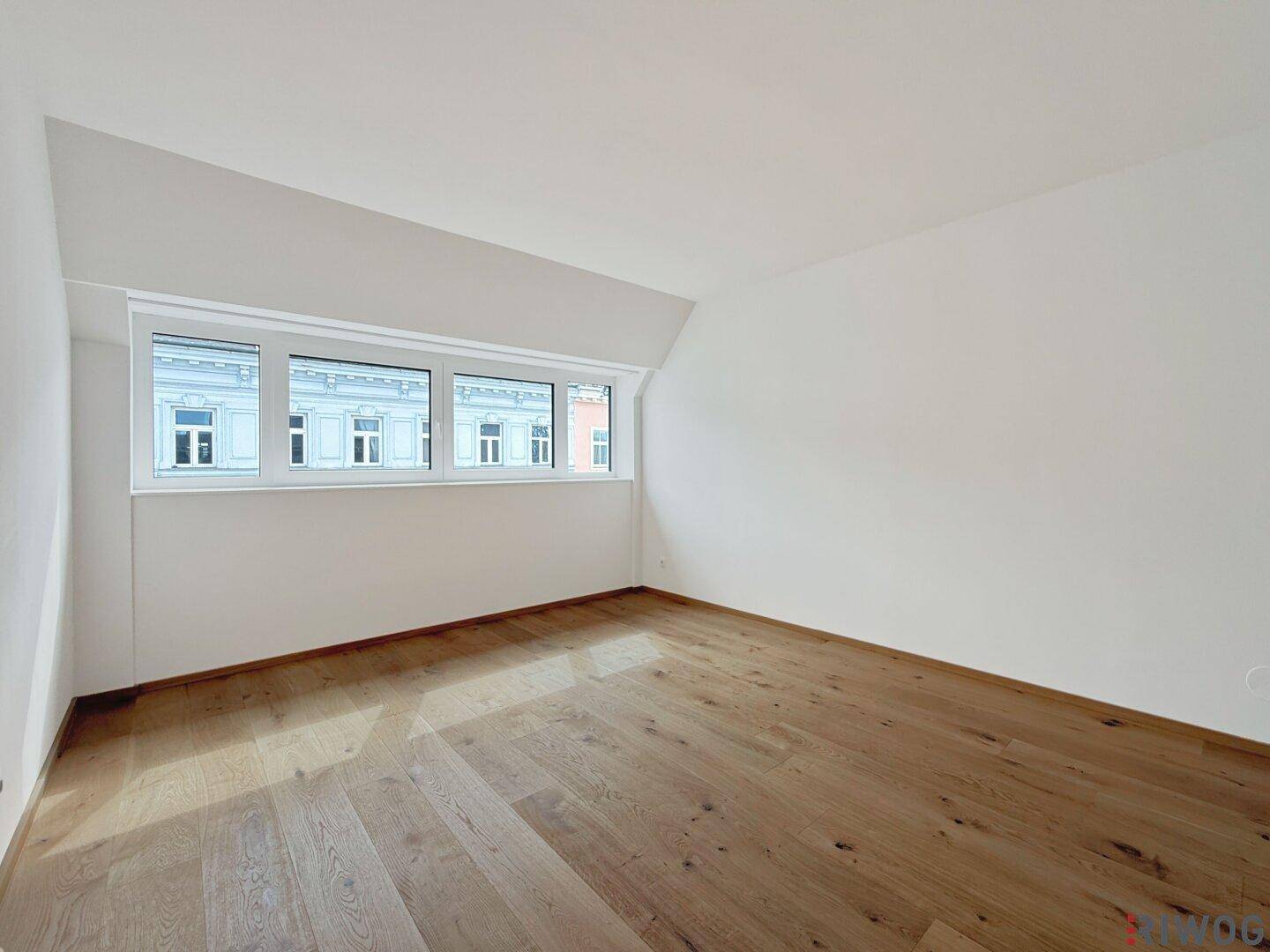 Geräumiges Zimmer mit Holzboden, breitem Fenster und Dachschräge, ideal als Schlaf- oder Arbeitszimmer.