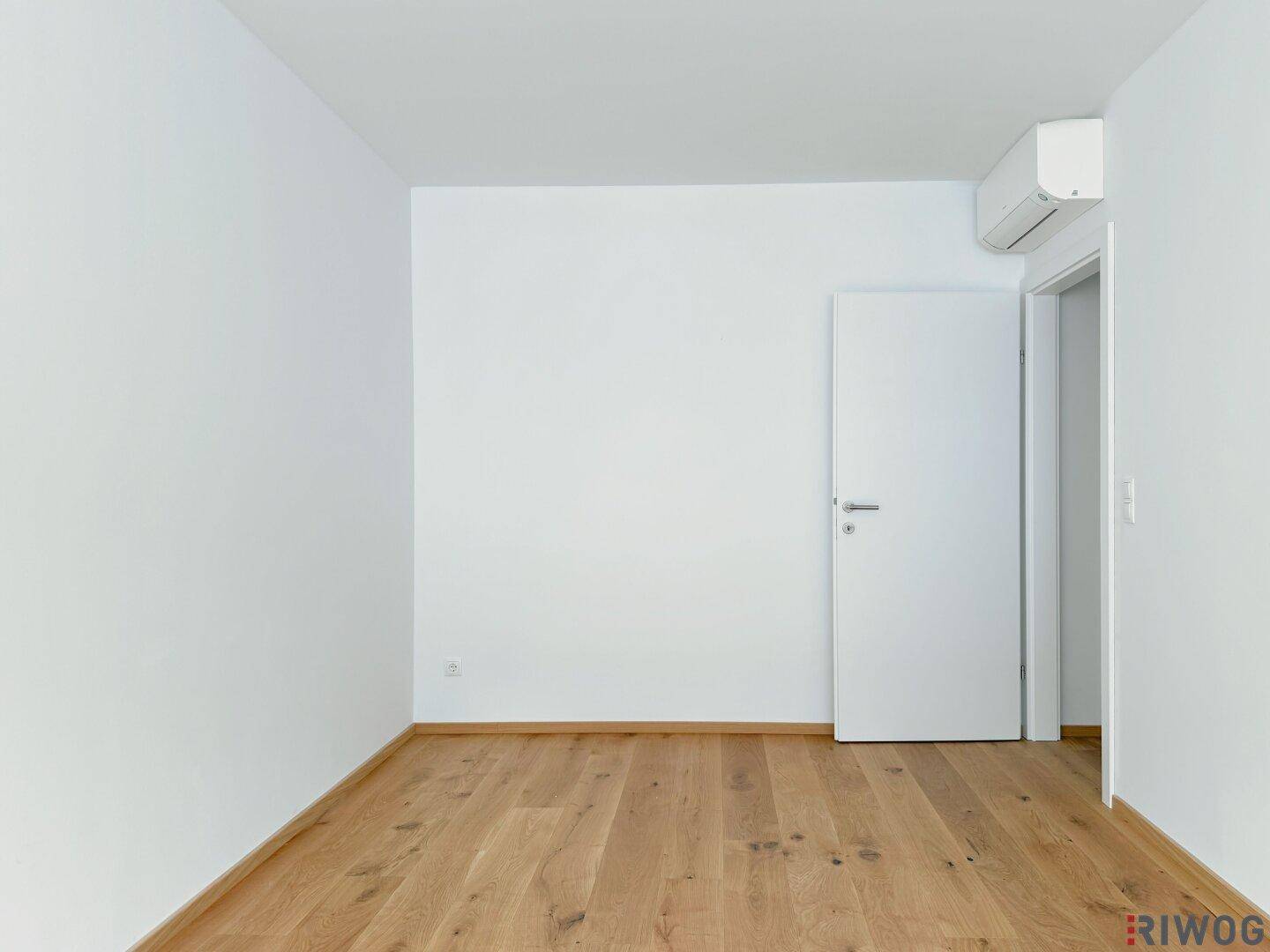 Leeres Zimmer mit Holzboden, offener Tür und Klimaanlage, bietet vielseitige Nutzungsmöglichkeiten.