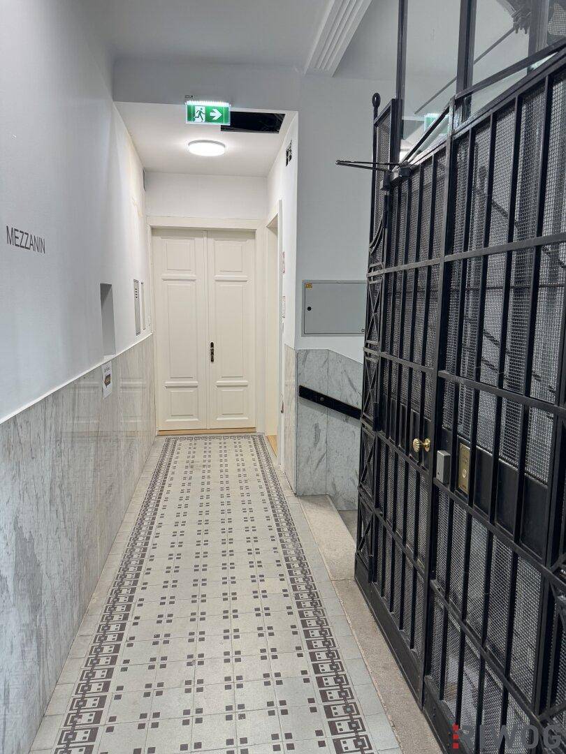 Heller Hausflur mit gemusterten Fliesen, Marmorverkleidung und historischem Aufzugsgitter.