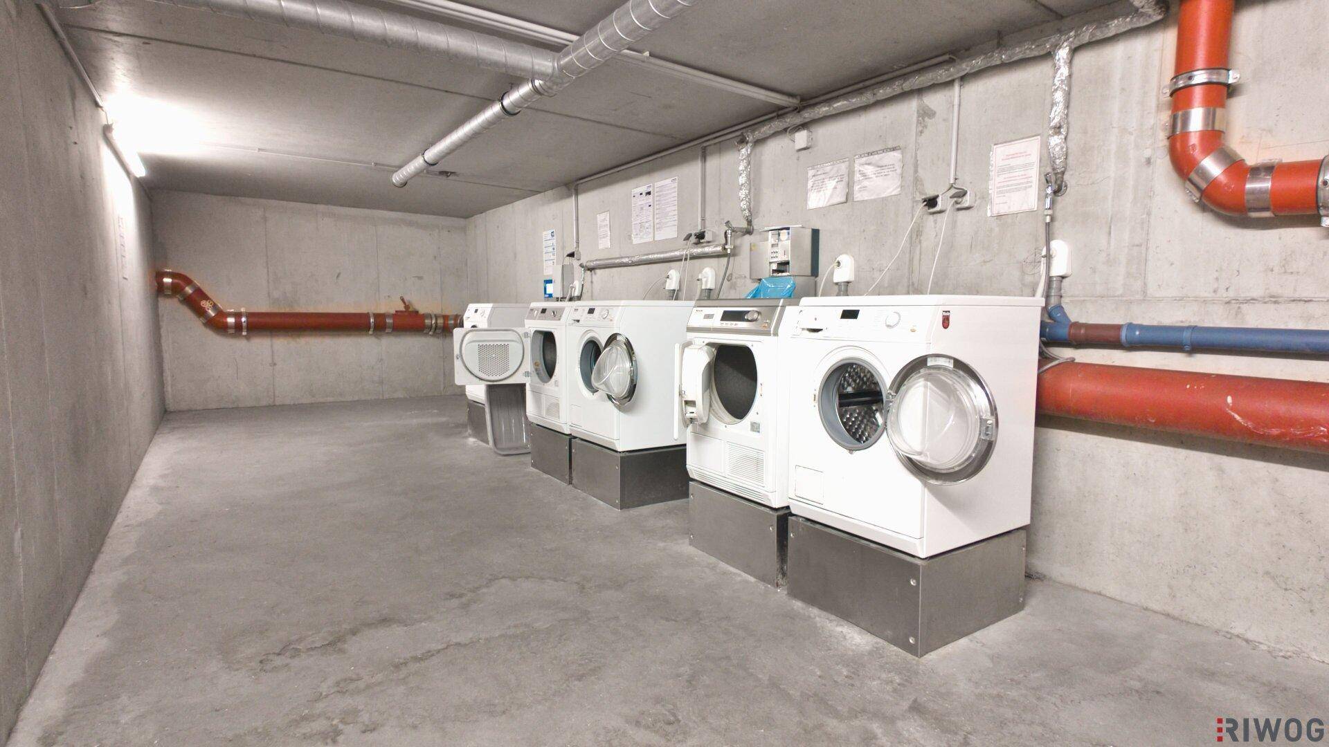 Funktionaler Waschraum im Untergeschoss mit mehreren Waschmaschinen und Trocknern sowie sichtbaren Leitungen.