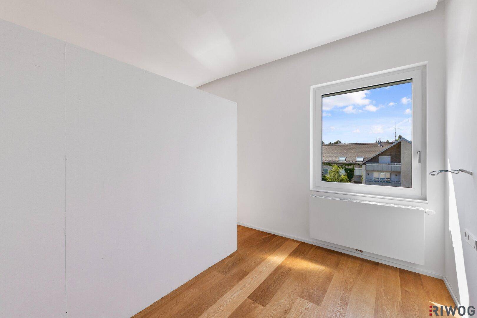 Leeres Schlafzimmer mit Holzboden, weißen Wänden und Ausblick auf Dächer und Bäume.