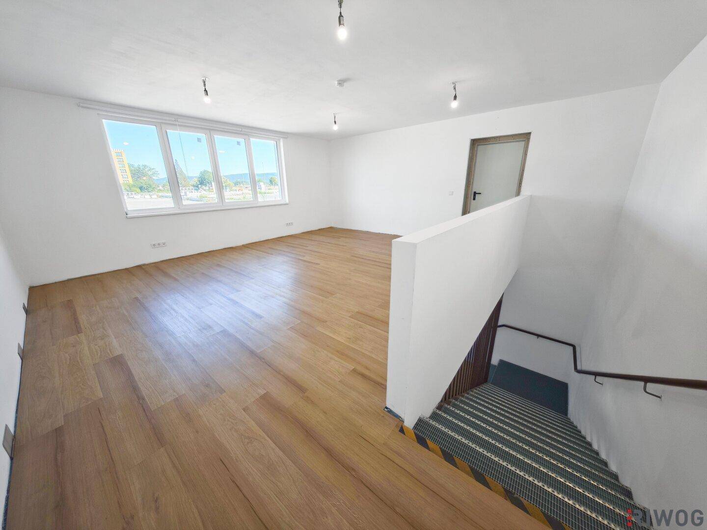 Großer, heller Büroraum mit Holzboden, großen Fenstern und einem Treppenabgang.