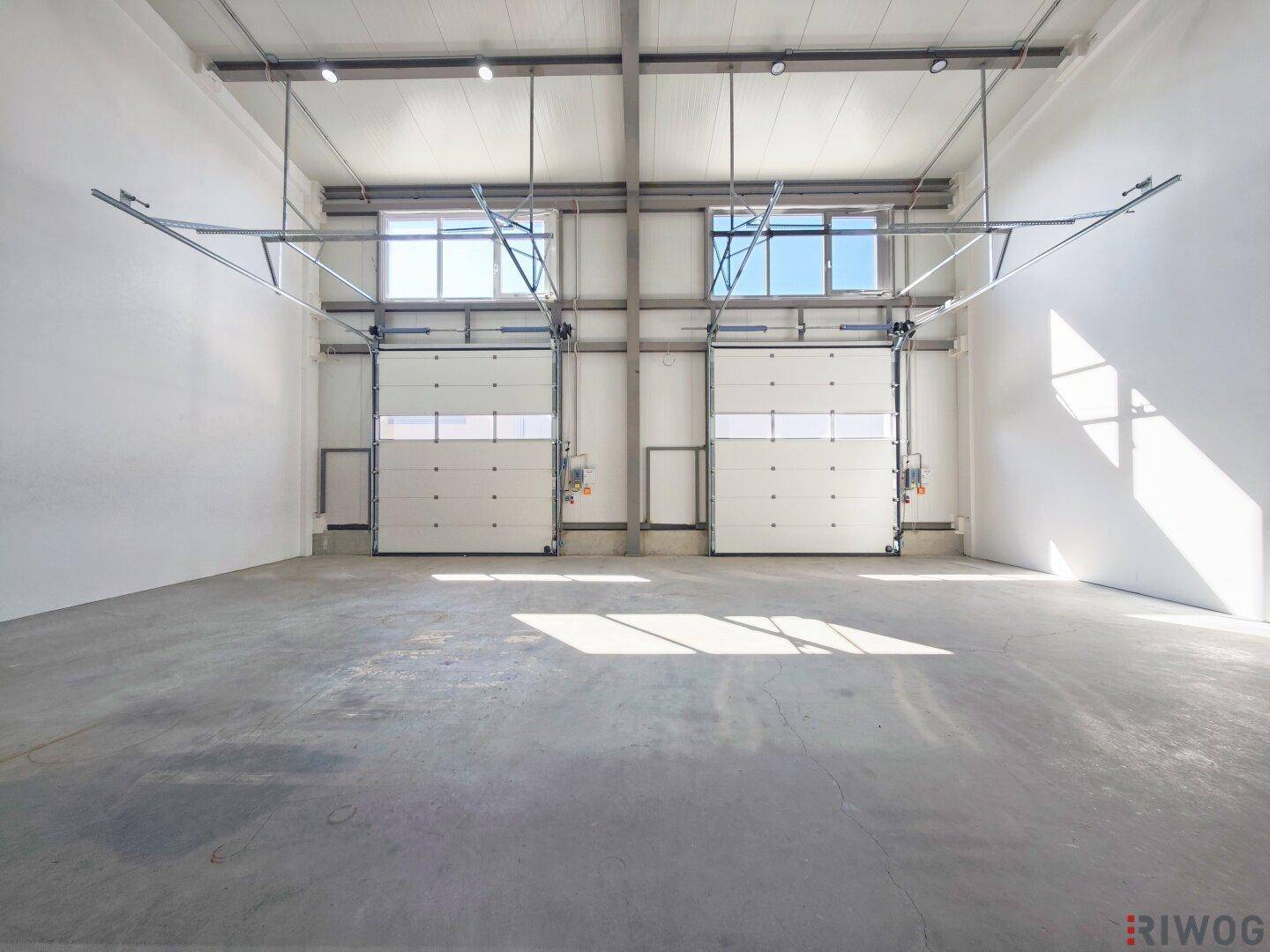 Innenansicht einer Lagerhalle mit zwei Rolltoren und Fenstern im oberen Bereich.