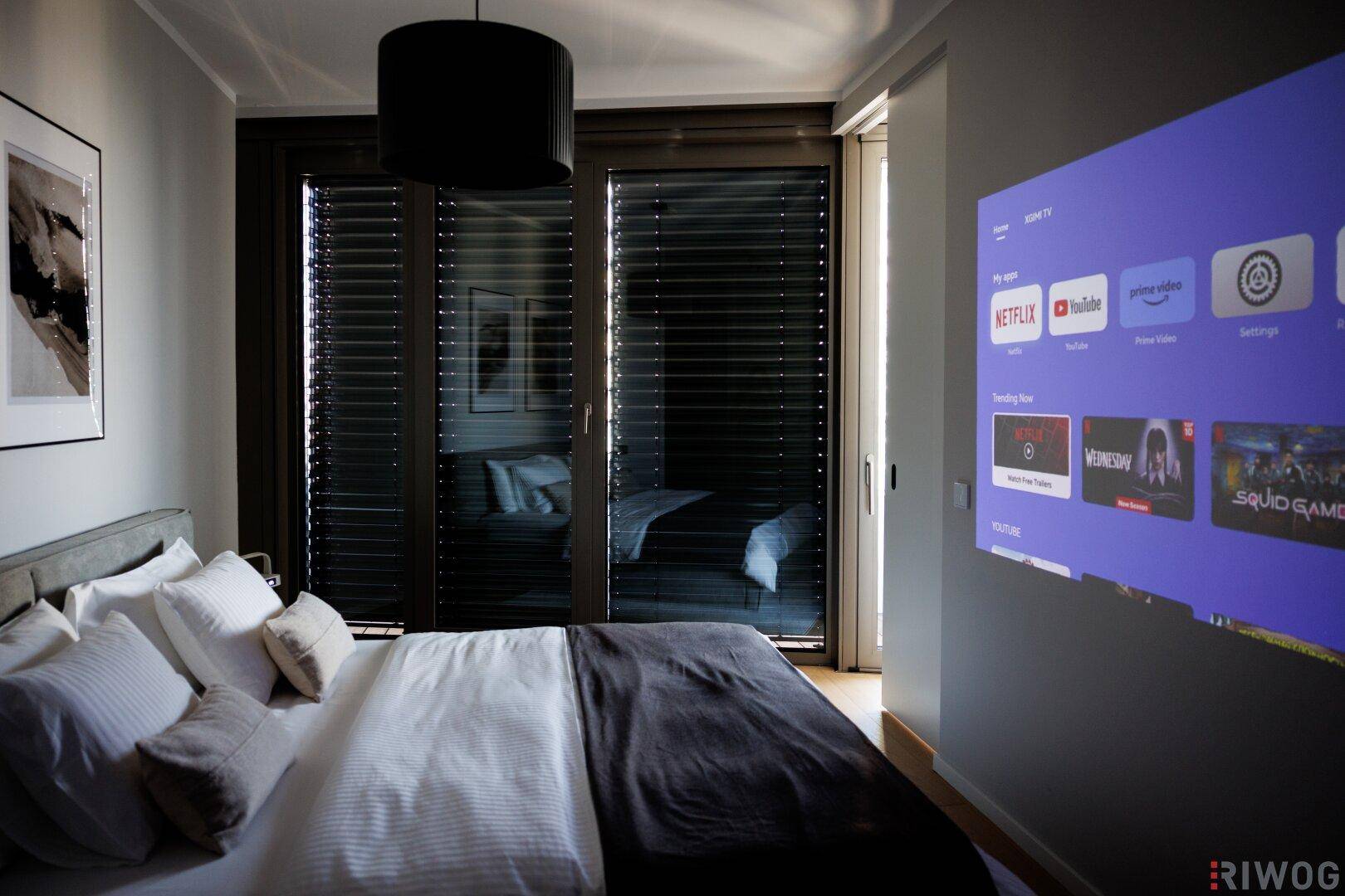 Schlafzimmer mit Projektor an der Wand, großem Bett und Zugang zu einem Balkon mit Jalousien.