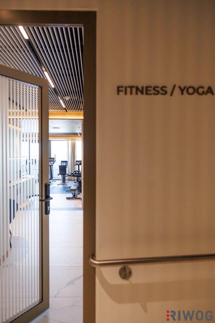 Geöffnete Tür zu einem modernen Fitness- und Yogabereich mit sichtbaren Trainingsgeräten im Hintergrund.