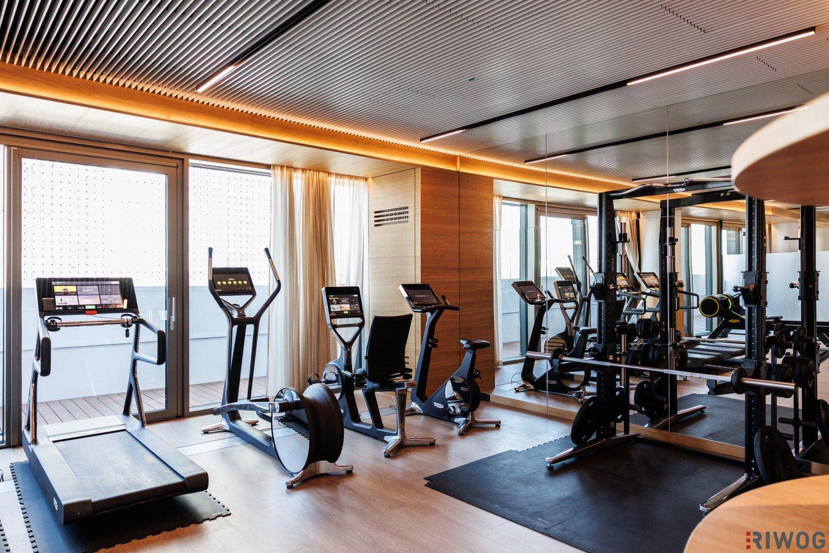 Geräumiger, heller Fitnessraum mit modernen Trainingsgeräten, Spiegelwänden und großen Fenstern.