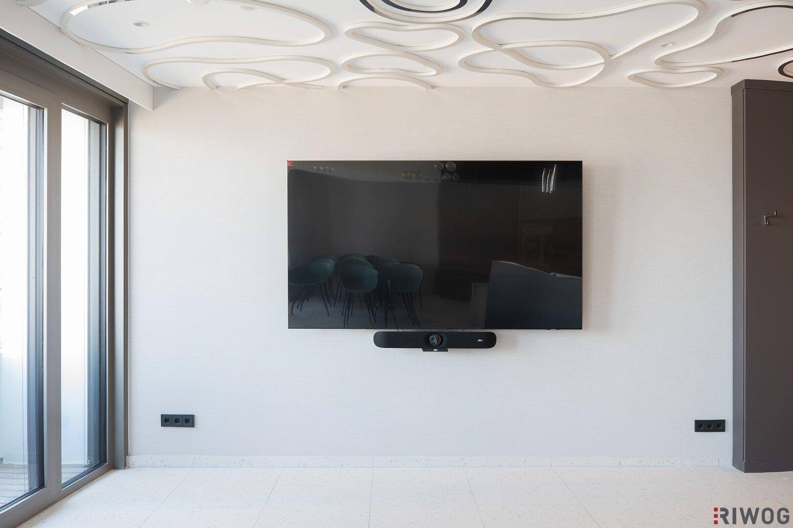 Heller Wohnbereich mit großem Wandfernseher und Zugang zu einem Balkon mit viel Tageslicht.