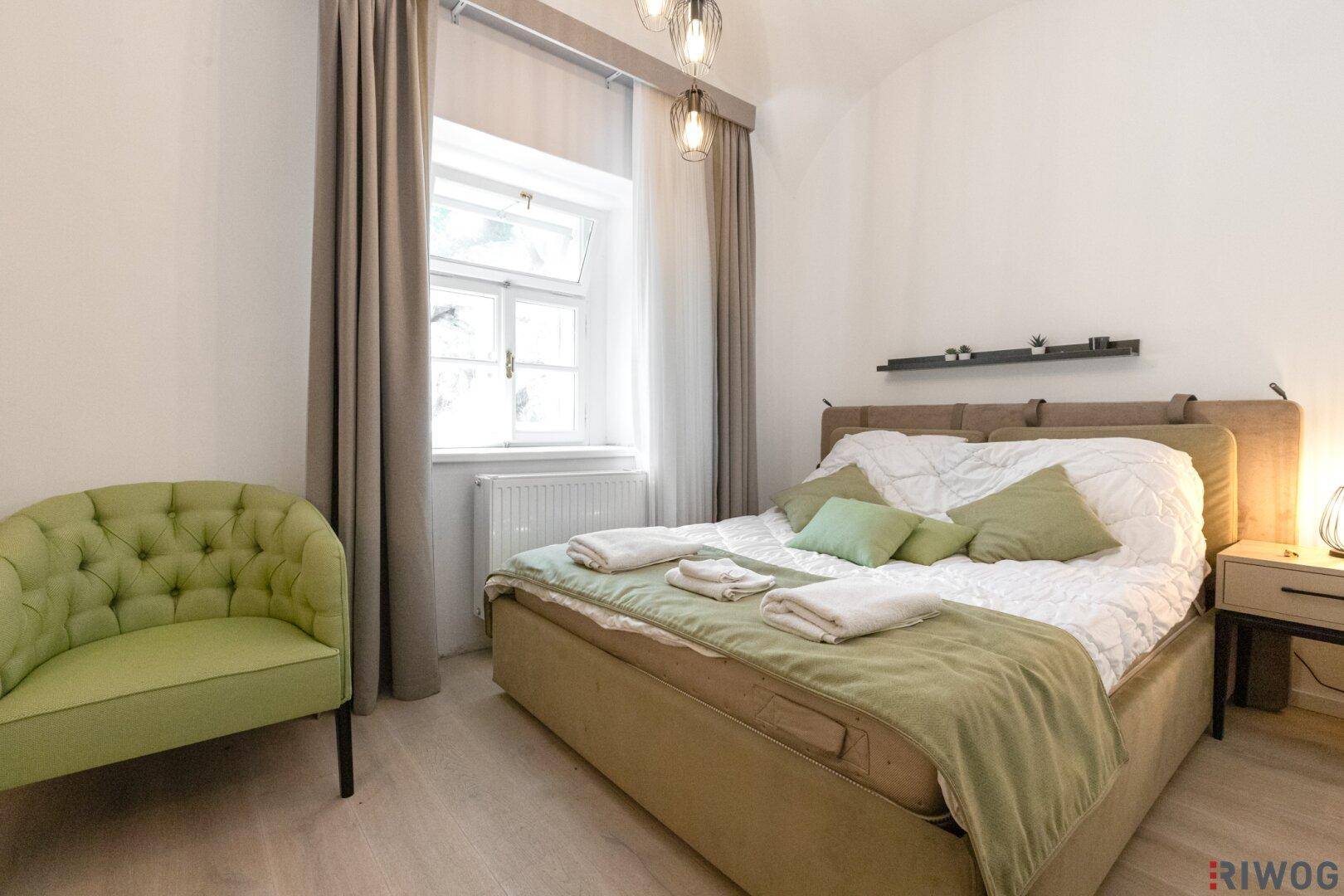 Helles Schlafzimmer mit Doppelbett, grünem Sessel und Fenster mit Vorhängen.