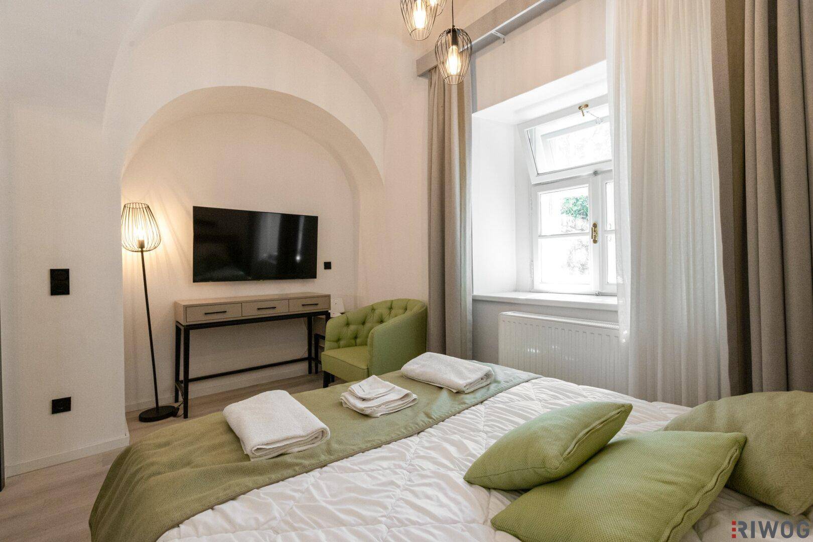 Gemütliches Schlafzimmer mit Doppelbett, Fernseher und einem großen Fenster für viel Tageslicht.