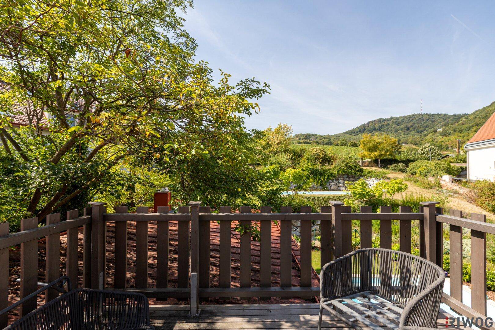 Sonniger Holzbalkon mit Sitzgelegenheiten und weitem Blick auf grüne Landschaft und Weinberge.