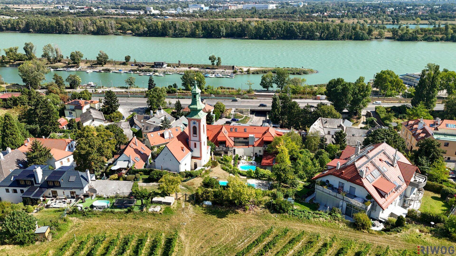Weitläufige Luftaufnahme eines Dorfes mit Kirche, Fluss, Weinbergen und grüner Umgebung.