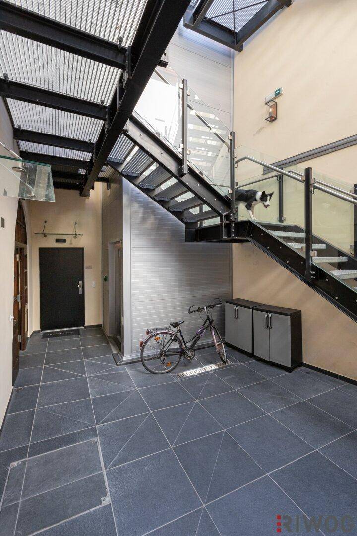 Moderner Eingangsbereich mit Treppe, Glasgeländer, Fliesenboden und Fahrradabstellplatz sowie Aufzug.